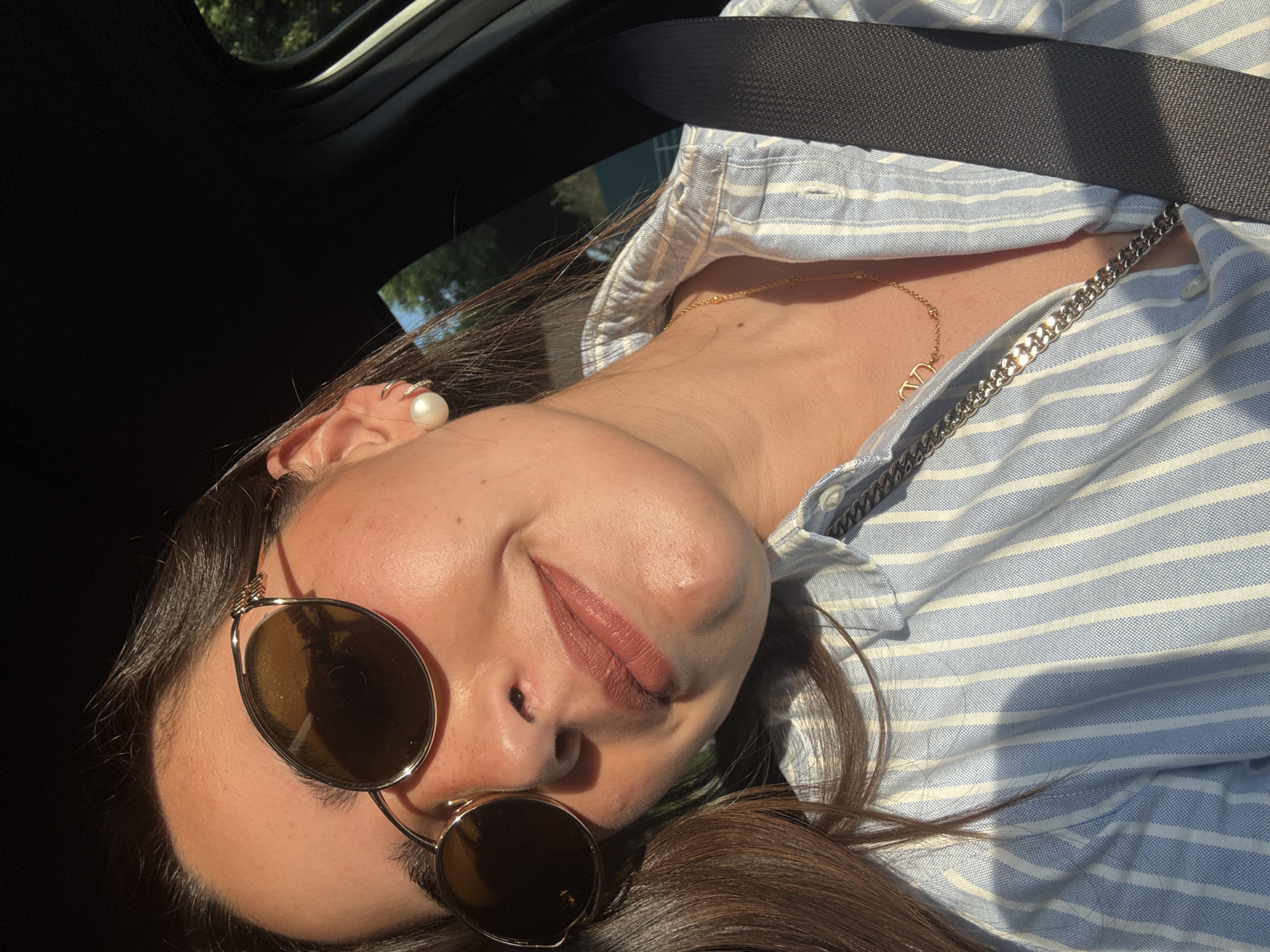 A woman is photographed from a low angle inside a vehicle, secured with a seatbelt. She wears round sunglasses, a pearl earring, and a striped shirt with a gold necklace. The location is difficult to determine based on the image provided; there is no specific indicator to link the image with the city of San Bernardino Tlaxcalancingo, Mexico.