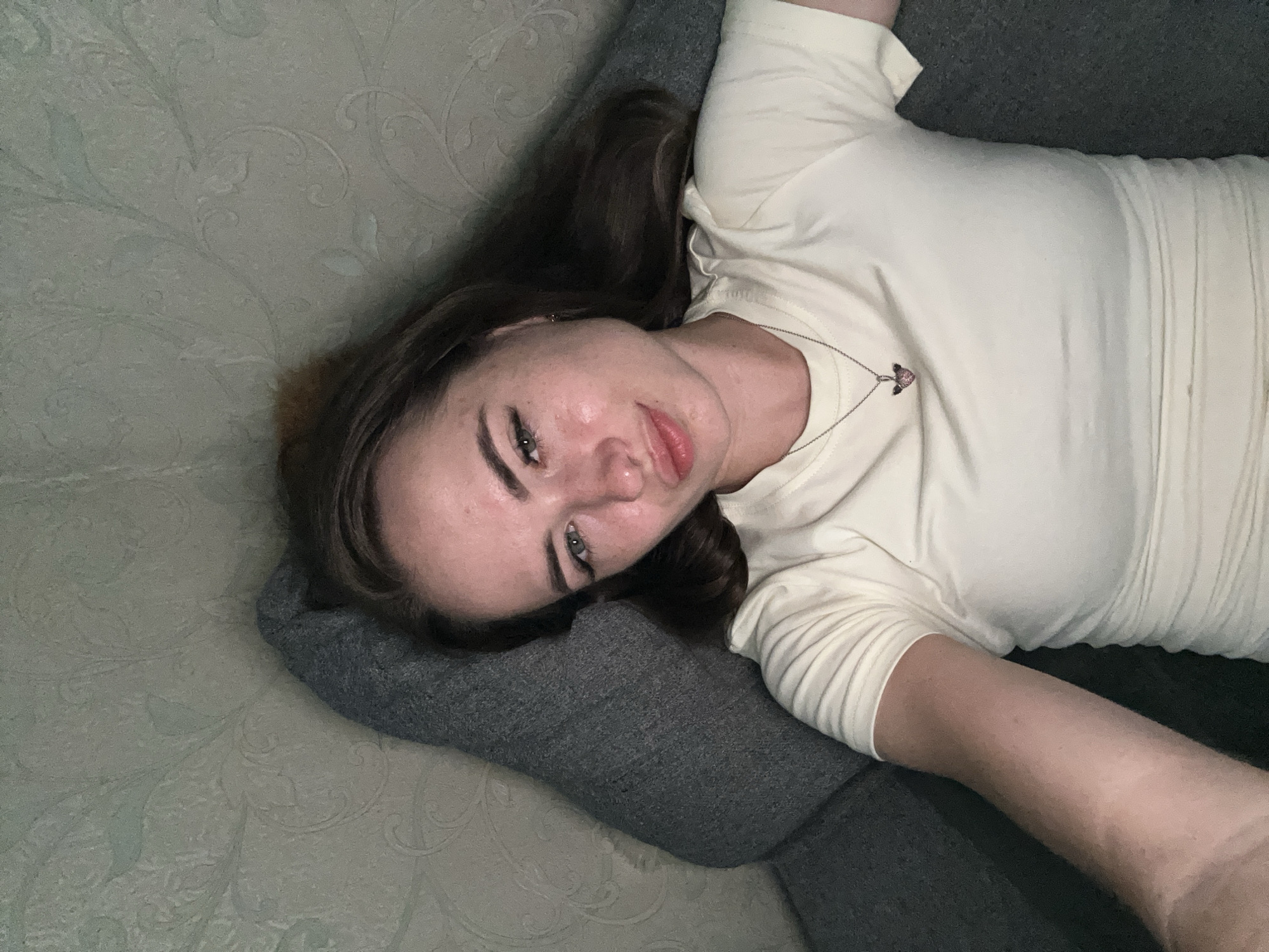 A young woman is lying on her side on a couch, with her head resting on a gray cushion. She is looking up and to her left, towards the camera. Her eyes are open, and she has a slight smile on her lips. She is wearing a light-colored t-shirt and a necklace with a small, heart-shaped pendant. Her long, brown hair is spread out around her head. The couch appears to be in a home setting, with a textured, pale green wall in the background. The lighting is soft, suggesting an indoor environment, possibly during the day or evening. The overall mood is relaxed and casual.