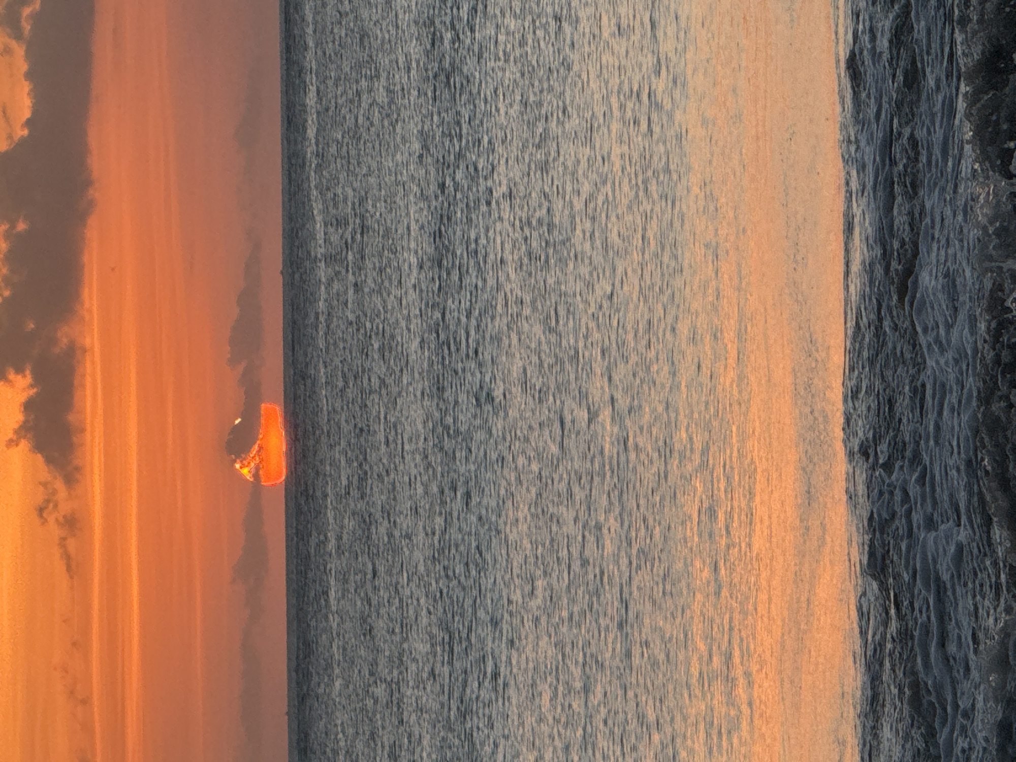 A vibrant orange sunset casts a warm glow over the ocean off the coast of Kuta Selatan, Indonesia. The sky is a gradient of oranges and pinks, with streaks of dark clouds. The sun, a bright orange orb, appears distorted as it dips below the horizon, partially submerged in the water. The ocean's surface is a deep blue, rippled by gentle waves. In the foreground, the waves are more pronounced, with white foam cresting and breaking onto the shore. The overall scene is serene and picturesque, capturing the beauty of a tropical sunset.