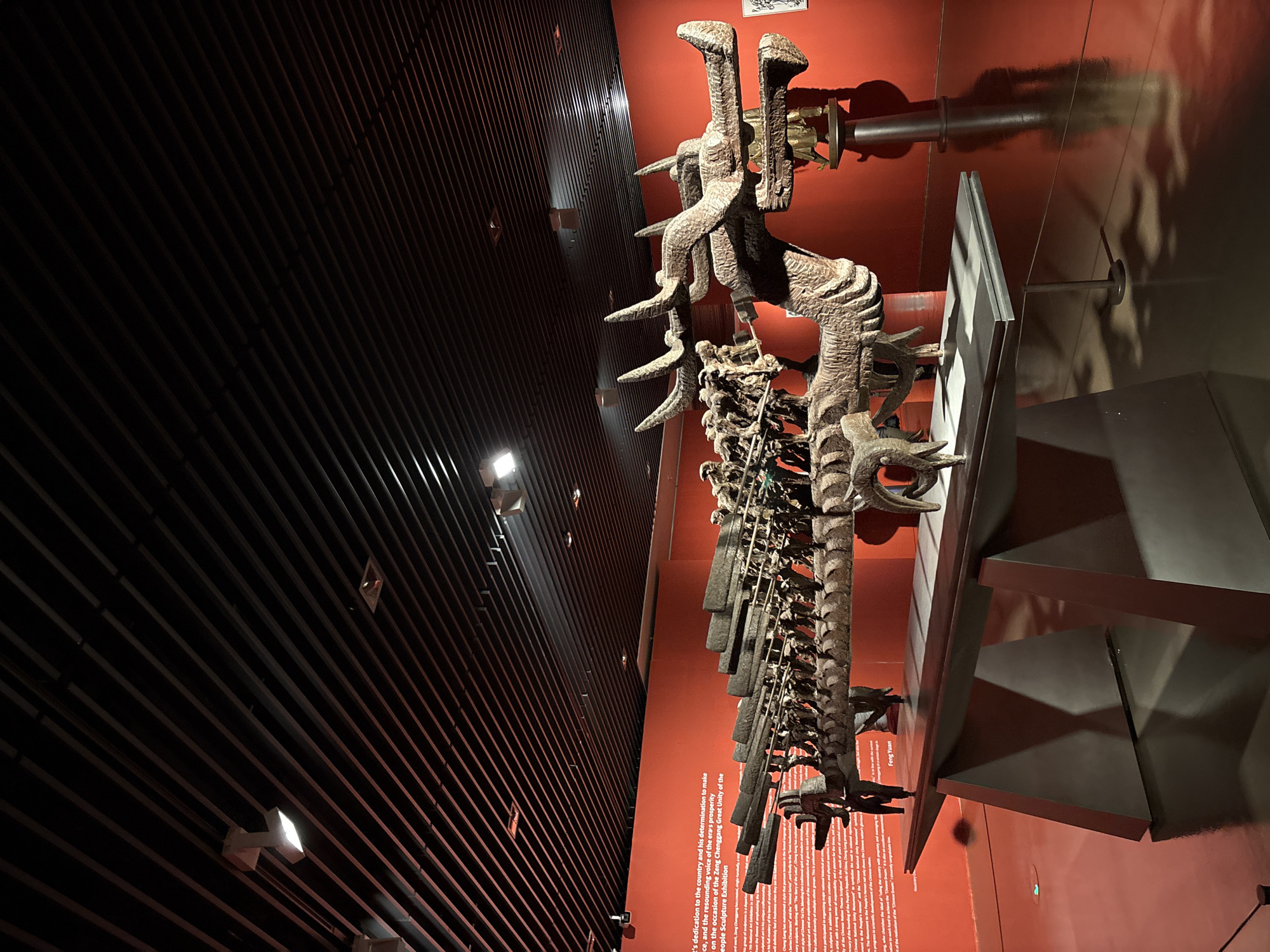 The image shows a large, elaborate dragon sculpture on display in a museum, with a bright red wall behind it. The sculpture appears to be made of wood or a similar material and is highly detailed, with intricate carvings and many segments. The dragon has a fearsome head with horns and a long, segmented body that extends across the frame. The sculpture is mounted on a grey metal stand. The museum interior has a modern design, with a dark, slatted ceiling and spotlights illuminating the exhibit. The floor is highly reflective, mirroring the sculpture and the surrounding environment. A portion of a descriptive plaque is visible to the left of the sculpture. The scene is well-lit, with the red background creating a dramatic contrast with the muted tones of the sculpture. The overall impression is one of a significant cultural artifact presented in a contemporary museum setting. The location is specified as Pudong, China, suggesting this is a cultural institution within that district.