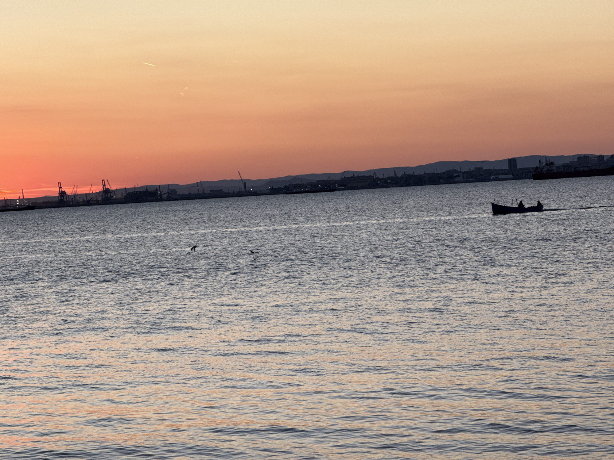 The image depicts a sunset over the Burgas Bay in Burgas, Bulgaria. The sky transitions from a pale yellow at the top to a vibrant orange and red near the horizon, suggesting the sun is setting. In the background, a silhouetted industrial port complex with cranes is visible along the coastline, and a range of hills or mountains forms the distant backdrop. The sea in the foreground is choppy, reflecting the colors of the sky. A small boat with at least two people is moving from left to right across the water, leaving a visible wake. Several birds are also present, some flying and others appearing to be on or near the water's surface. The overall scene captures the natural beauty of the sunset juxtaposed with the industrial landscape of the port.