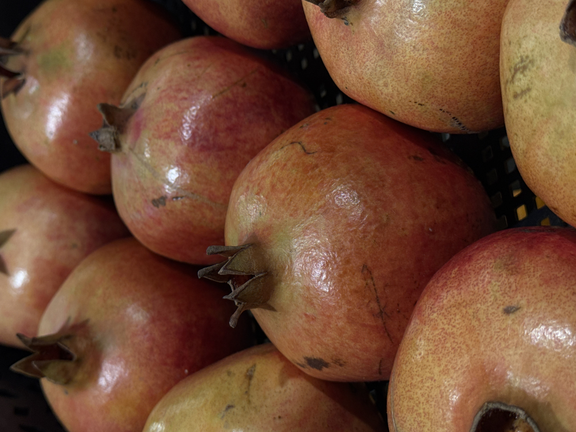 This media file is a close-up, eye-level shot of a pile of pomegranates. The fruits are the primary subjects, filling the entire frame. They are predominantly a warm, reddish-orange color with some showing hints of green and yellow. The texture of their skin is visible, appearing slightly leathery and dotted. The pomegranates are stacked in a way that suggests they are being displayed or stored, possibly in a market or grocery store setting.
The lighting appears to be artificial, casting a gentle glow on the fruits and highlighting their smooth surfaces. There are no people visible in the image. The background is dark and out of focus, with hints of a dark, possibly black, mesh or grate structure behind the pomegranates, which could indicate a basket or crate.
There are no specific cues about the time of day or weather. The setting, while likely indoors in a place where produce is sold, is not explicitly identifiable beyond that. No text is visible in the image. The overall impression is one of abundance and freshness, with the focus solely on the natural beauty of the pomegranates. The image is static, with no discernible activity taking place. The location context of Moscow, Russia, does not provide any specific visual cues within the image itself.