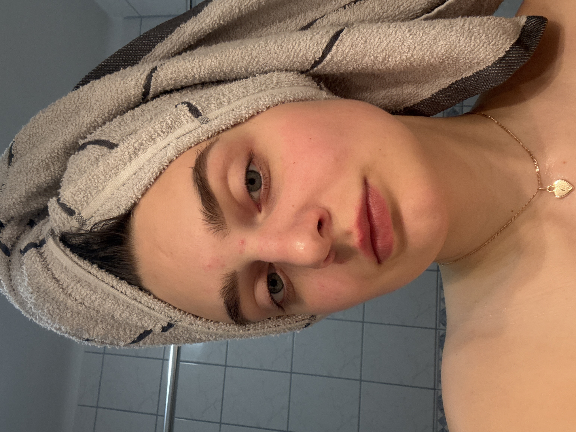 A young woman with a towel wrapped around her head poses for a close-up photograph in a bathroom. She has light skin, blue-grey eyes, and full lips. A gold necklace with a heart-shaped pendant is visible around her neck. The bathroom features tiled walls, with tiles visible in the background. The location cannot be determined definitively as Hainburg an der Donau, Austria from the image.