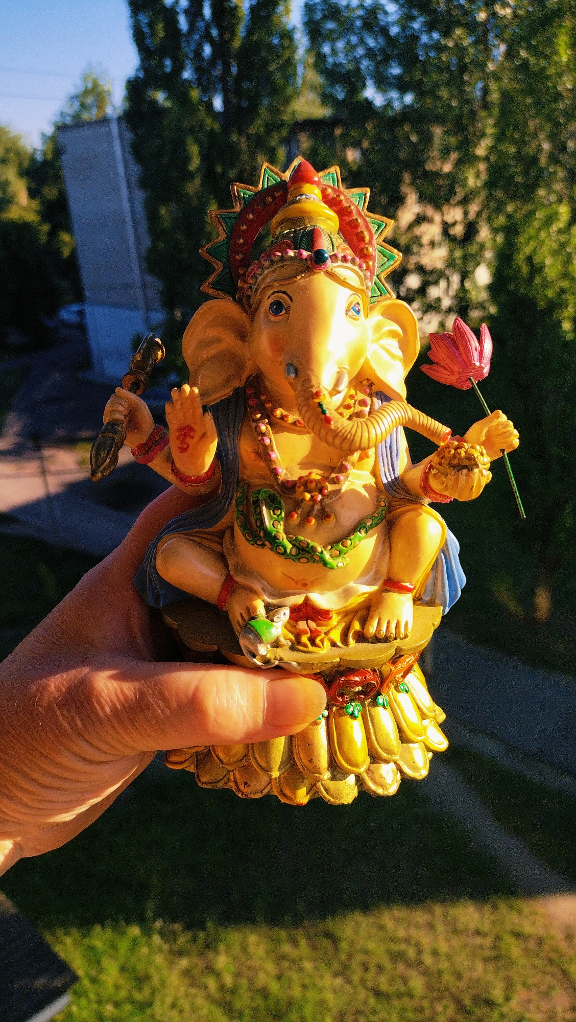 A hand holds a golden statue of Ganesha, a deity from Hinduism, in front of a blurred background of green trees and a building. The statue is adorned with colorful jewelry and holds a lotus flower in one hand and a modak (sweet) in another. The image appears to be taken outdoors during daylight, with sunlight casting shadows on the statue and the hand. The background suggests an urban environment, possibly a residential area, given the presence of a building and trees. The overall impression is one of a spiritual or decorative object being displayed in an outdoor setting. The location of Horishni Plavni, Ukraine is not identifiable from the image itself, as there are no specific landmarks or contextual clues to indicate this location.