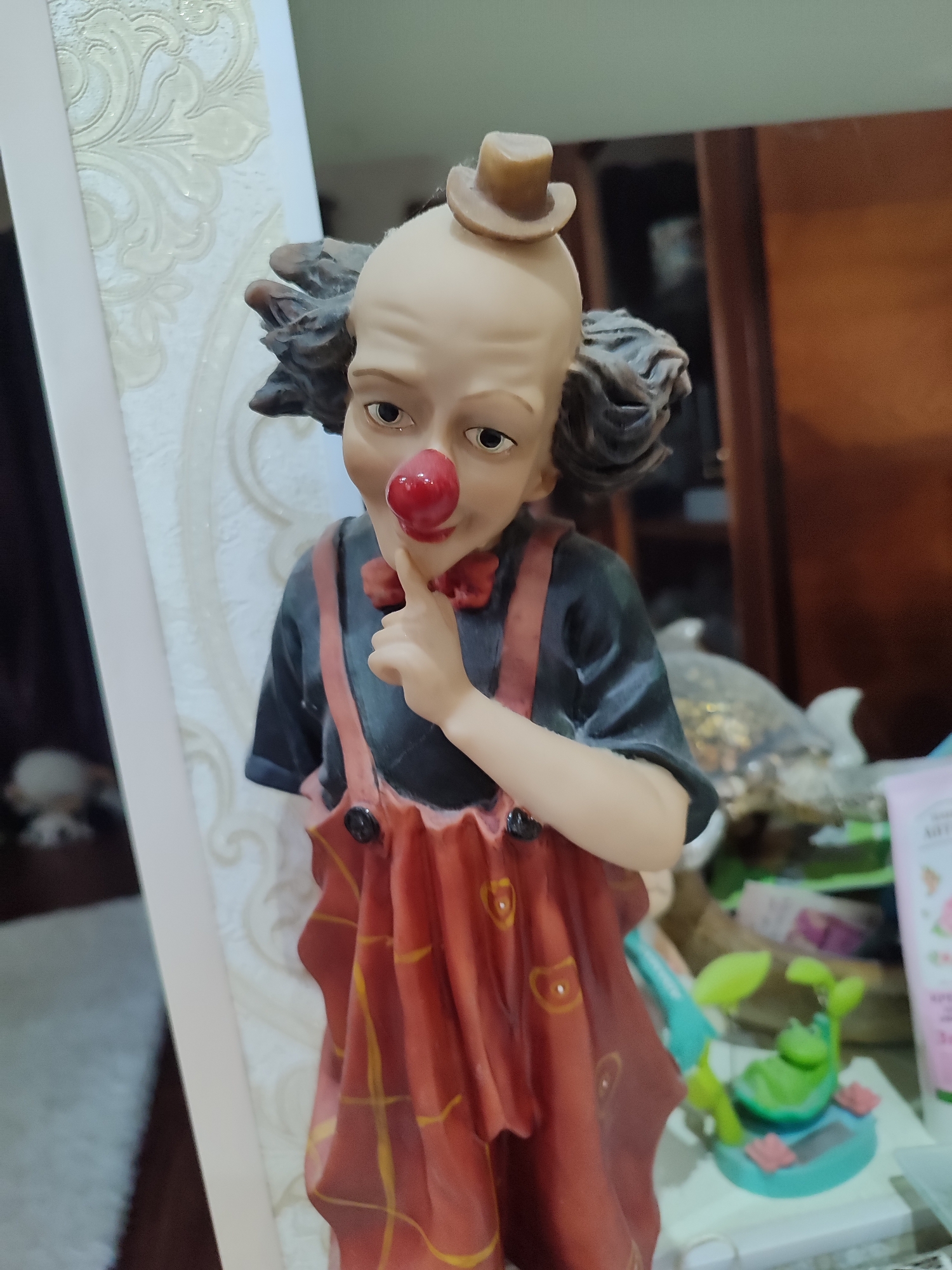 This is a close-up, eye-level, medium shot of a porcelain figurine of a clown. The clown has a bald head with tufts of dark, curly hair, a large red nose, and a small brown top hat. The clown is wearing a black shirt with red suspenders and a red, ruffled skirt with a yellow plaid pattern. Its right index finger is raised to its lips, suggesting a gesture of secrecy or silence. The figurine stands on a white surface, possibly a shelf or table, with a decorative textured wallpaper visible to the left. In the background, there are blurred objects, including what appears to be a collection of decorative items, possibly including a turtle figurine, and a tube of what seems to be a cosmetic product with visible pink and green branding. The overall lighting is warm and appears to be from an indoor source. The setting is likely a home environment, potentially a living room or display area. There is no discernible text visible on the figurine itself.