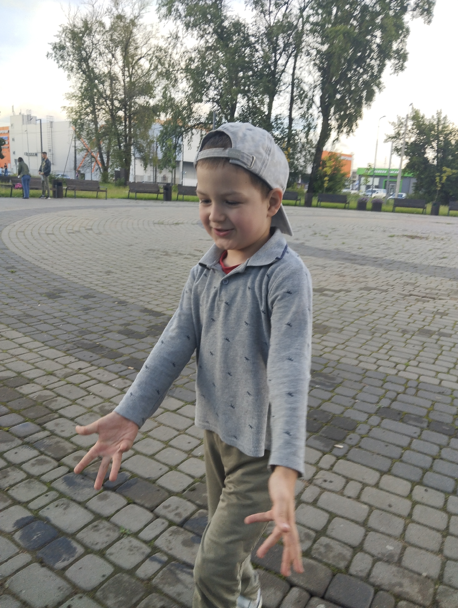 A young boy wearing a grey baseball cap and a grey long-sleeved shirt with a pattern of small dark figures is standing in a paved outdoor area with his arms outstretched, palms up. He is looking to his left, and his mouth is slightly open, suggesting he might be speaking or laughing. The ground is covered with interlocking grey pavers. In the background, trees with green foliage are visible, along with some buildings and park benches. The sky is overcast. The location is Kingisepp, Russia.