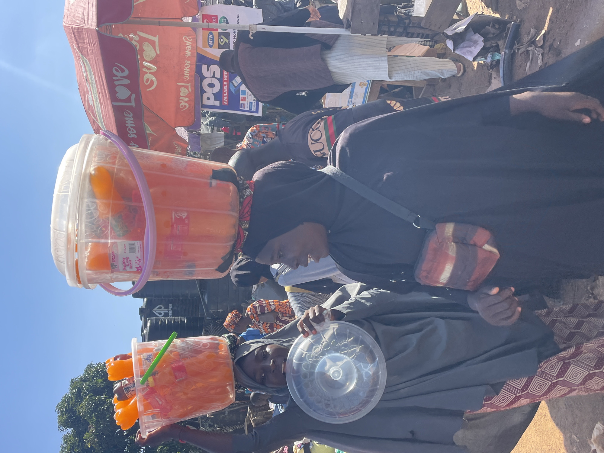 The image shows a street scene in Gura Top, Nigeria, on a sunny day. Two women are the primary subjects. The woman on the left, wearing a gray hijab and dress, balances a large clear plastic bucket filled with orange-colored liquid and bottles on her head. She is also holding a clear plastic basin in front of her. The woman on the right, wearing a black hijab and dress, has a similar bucket on her head and is looking to her left. She has a small, multi-colored bag slung across her chest.
In the background, other people are visible, some in traditional attire. Various stalls and umbrellas are set up, including one with the text "some Love" and a POS advertising banner. The ground is dusty and uneven, indicative of a marketplace. Trees are visible in the far background under a clear blue sky. The overall impression is a busy, outdoor commercial environment.