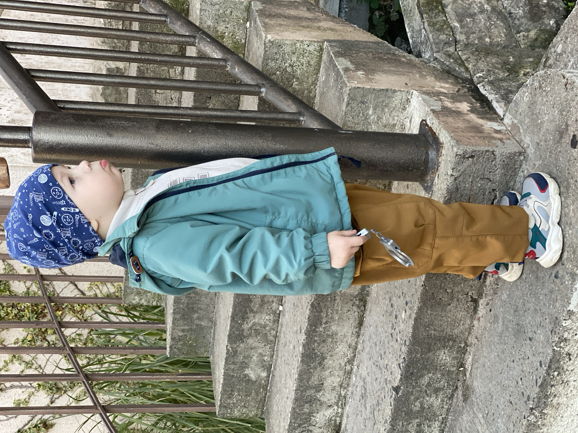 A young child in a blue space-themed cap, a light blue jacket, and brown pants stands on concrete steps next to a metal railing. The child is holding a small, metallic object in their right hand. The background includes concrete structures and some greenery. The image appears to be taken outdoors. Based on visual context, it is not possible to verify it is in Sochi, Russia.
