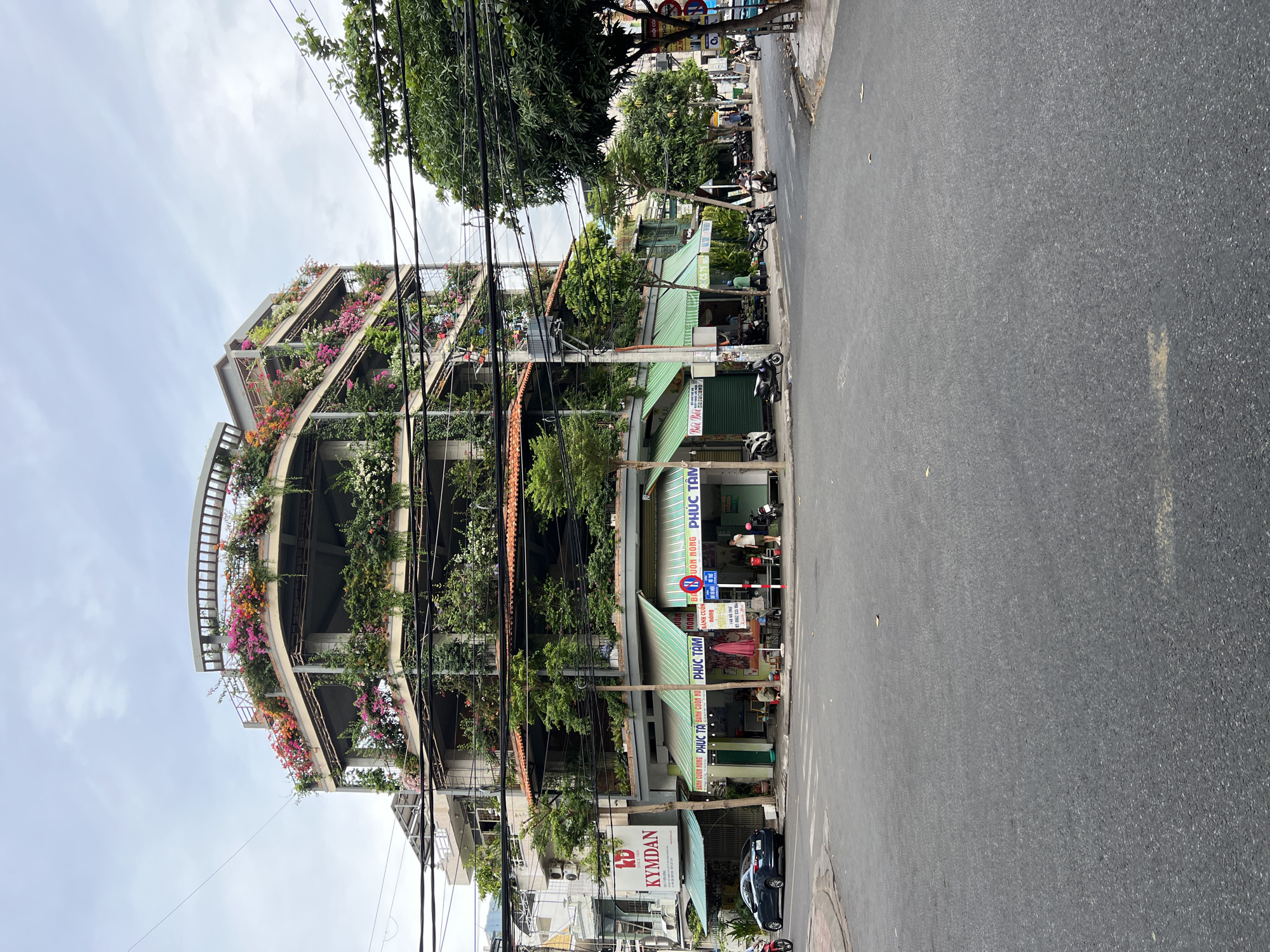 This image depicts a multistory building in Nha Trang, Vietnam, characterized by extensive vertical gardens and floral displays on multiple levels. The building features open-air levels with concrete columns and railings, adorned with a profusion of green foliage and vibrant flowers, particularly bougainvillea in shades of pink, purple, and orange. The ground floor of the building houses several businesses with green striped awnings, including "PHUC TAM" and "KYMDAN," signage indicating it is a mattress store. A dark blue sedan is parked on the street in front of the building, and several motorcycles are visible parked on the sidewalk and street. Two individuals are present near the businesses on the ground floor, one appearing to be a shopkeeper and the other a customer. The street is paved, and overhead power lines crisscross the sky, typical of urban Vietnamese streetscapes. The environment appears to be an intersection in a residential or commercial district under an overcast sky.