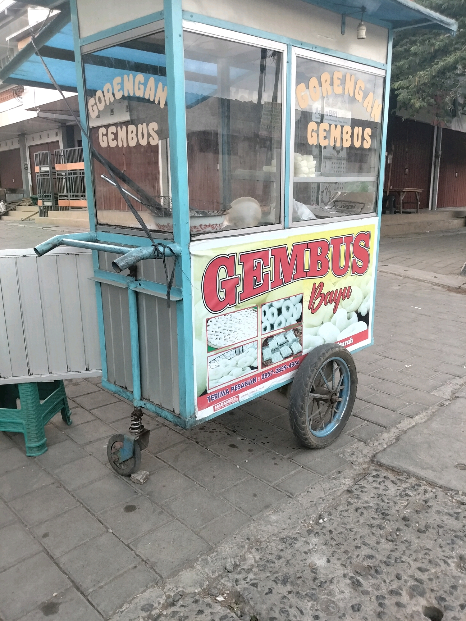 The image shows a food cart for sale in Pituruh, Indonesia. The cart is blue and white, with a sign that says "GORENGAN GEMBUS." The cart is parked on a sidewalk, with a street in the background. There are buildings on either side of the street, and trees in the distance. The cart appears to be empty, and there are no people in the image. The sign on the cart also displays a phone number for orders: 0857-2859-4866. The cart has one large wheel on the right side, and a smaller, swivel wheel on the left side. There is a foldable table attached to the cart on the left side.