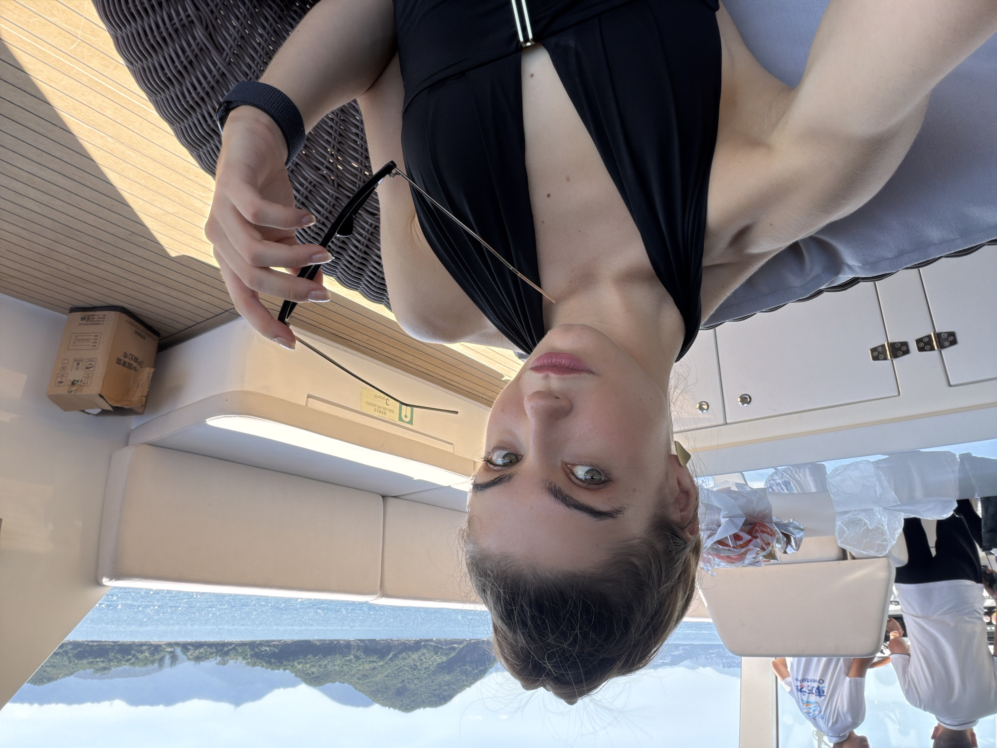 A young woman with her hair pulled up in a bun takes a selfie on the deck of a yacht. She is wearing a black halter-neck swimsuit and holding a pair of sunglasses. The yacht is at sea, with a view of green, hilly islands in the background. The deck is made of wooden planks, and there are white cushioned seats. In the background, two men in white shirts are visible near the helm of the yacht. The water appears blue and calm under a clear, sunny sky. The scene is set in Hong Kong, China, likely in the outer islands or harbors.