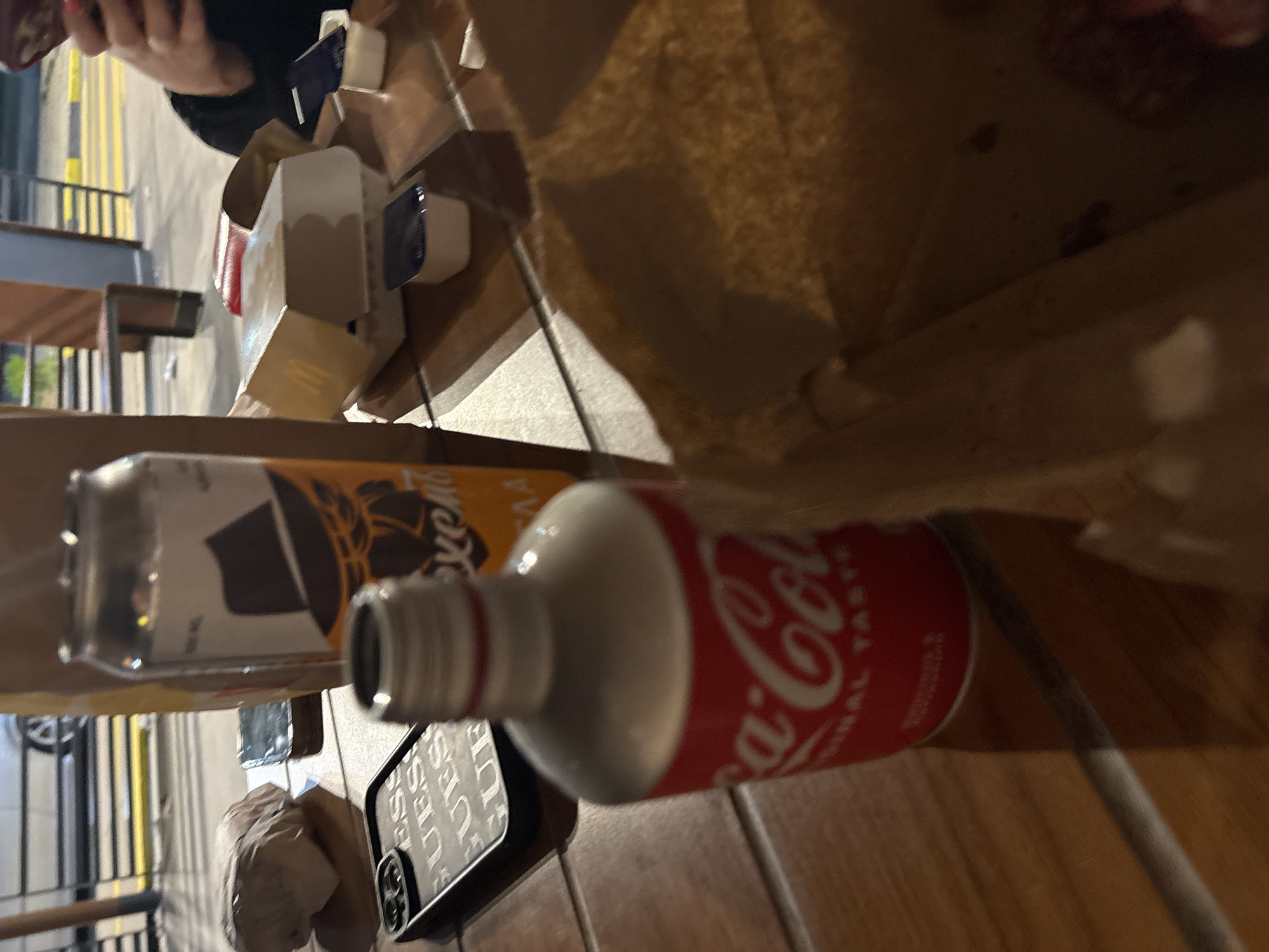 The image shows a still life on a wooden table, featuring a Coca-Cola glass bottle with a red and white label, a cardboard box from a food establishment, a black smartphone, and condiments. The scene appears to be set in a restaurant or outdoor dining area, evidenced by the glimpse of a street with yellow lines and metal railings in the background. A person's hand holding the box is visible. Based on the information provided, it is not possible to definitively confirm whether the location is Plovdiv, Bulgaria.
