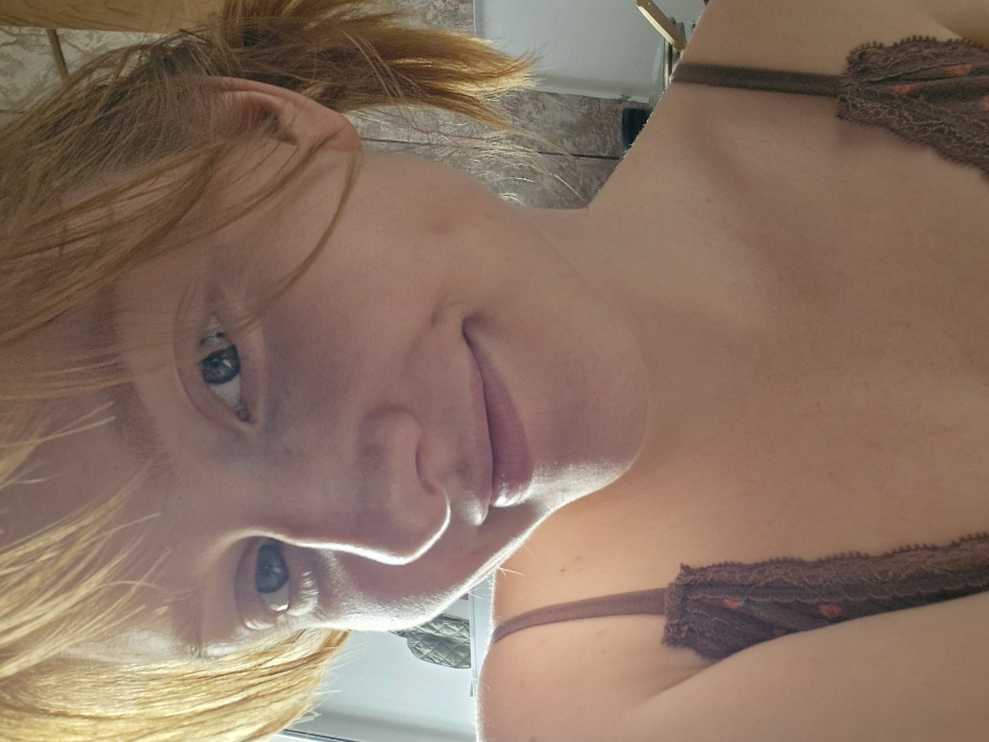 A close-up, tilted view depicts a fair-skinned individual with red hair pulled back in a ponytail. The person is wearing a brown, lacy bra. The background appears to be a tiled wall, possibly a bathroom. The setting is not identifiable as being in Yekaterinburg, Russia.
