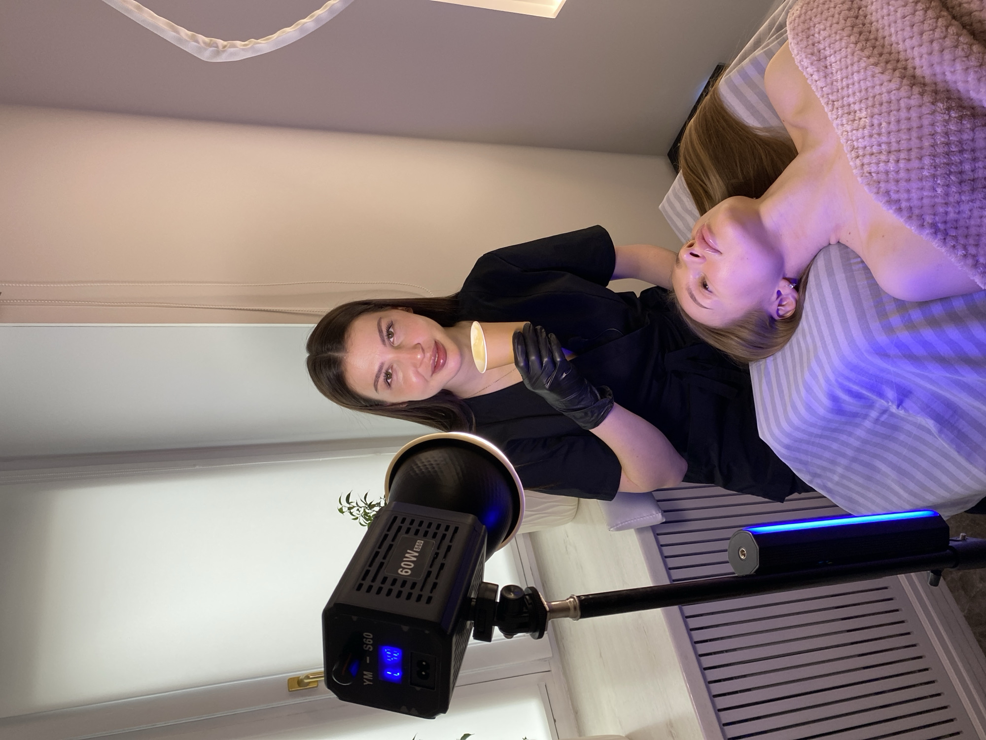 The image captures an indoor scene, likely a beauty salon or treatment room, well-lit by artificial sources. Two women are the central subjects.
On the left side of the frame, a woman, appearing to be a technician or aesthetician, stands or sits upright. She has dark hair, wears a black uniform top and black gloves. She holds a small, light brown cup containing a light yellow, creamy substance, possibly wax or a cosmetic product. She looks directly at the camera with a pleasant, slight smile.
To her right, a second woman, the client, lies on her back on a treatment bed. Her blonde or light brown hair is spread out on a striped pillow. A light pink or beige textured blanket covers her torso. Her head is turned slightly towards the left, and her skin, particularly on her neck and chest, shows a distinct purple-blue tint from a specialized light source. She appears relaxed and comfortable.
Prominently featured in the lower part of the frame are two pieces of lighting equipment. On the left, a large, black LED light unit is mounted on a stand, displaying text such as "60W" and "YM - S60," along with a digital readout of "U 4.0." To its right, a long, cylindrical black light bar emits a vibrant blue light. In the background, a small plant in a white pot and a white slatted structure are partially visible.
The scene suggests a beauty or aesthetic treatment is underway, possibly involving hair removal, a facial, or a similar cosmetic procedure, given the specialized lighting and products. The image is oriented sideways, with the technician appearing vertical and the client horizontal relative to the frame. The ambiance is professional and serene.