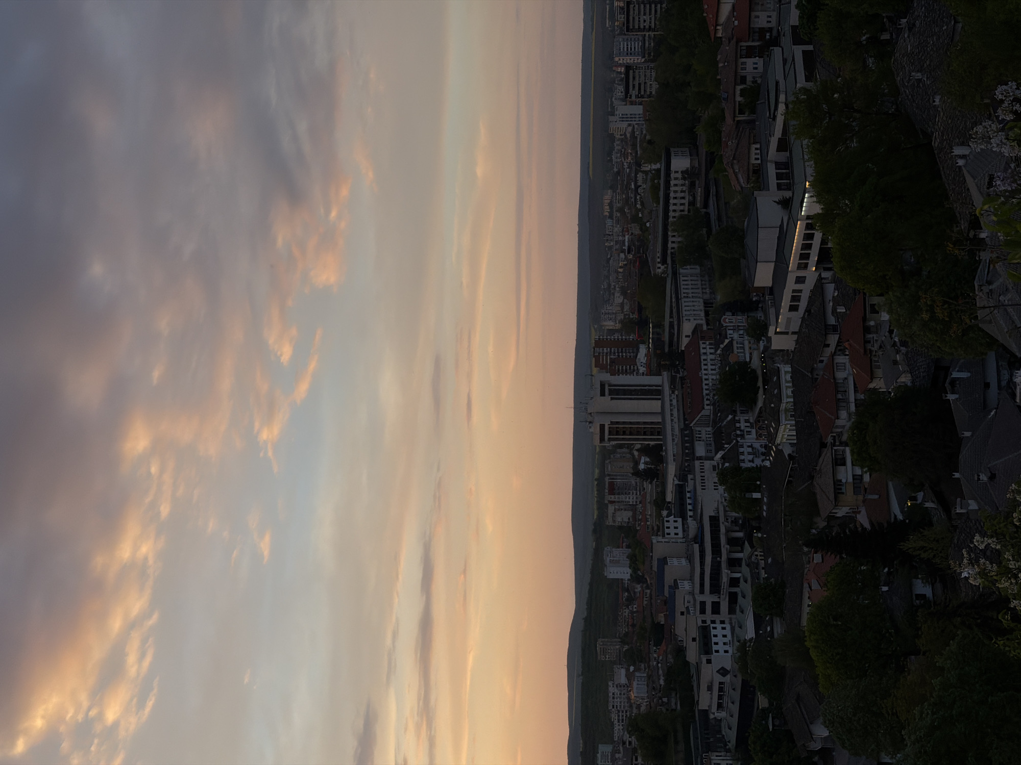 The image shows a cityscape under a colorful sky at sunset. On the right side is the city of Lovech, Bulgaria, with buildings of varying sizes and architectural styles. There are residential buildings, high-rise apartments, and commercial structures among the dark roofs and greenery of trees. The light fades towards the foreground, making it difficult to make out specific details. On the left side is a light-colored sky, illuminated with pink and orange hues indicative of sunset. The sky dominates the left half of the image, with scattered clouds creating an atmospheric effect.