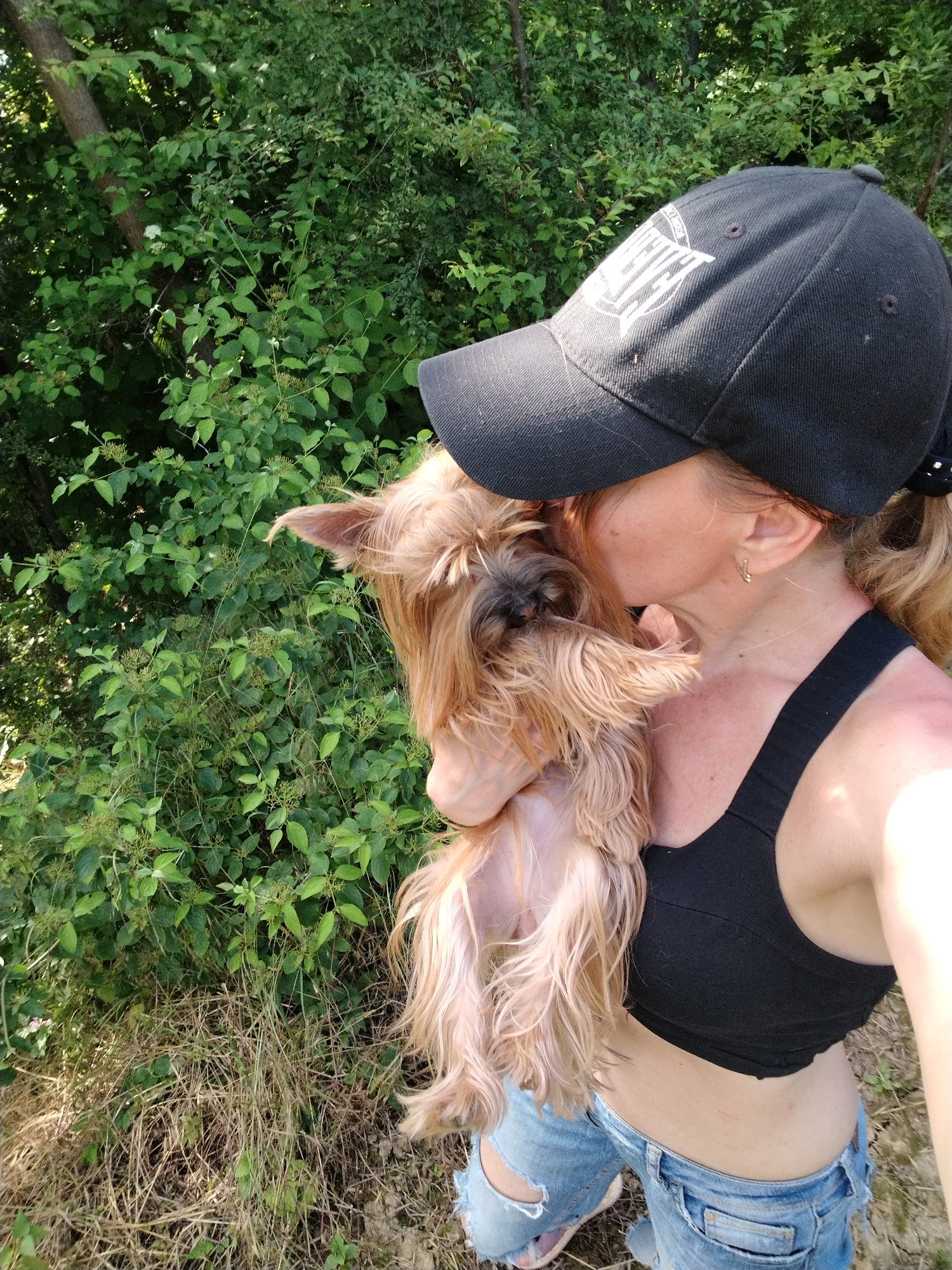 This image, captured in Novobzhegokay, Russia, displays a female individual holding a small dog in an outdoor, natural environment. The woman, whose face is largely obscured by a black baseball cap and the dog's head, is positioned with her right side visible to the viewer. She wears a black baseball cap with a white emblem on the front, a black cropped top or sports bra, and light blue distressed jeans with visible tears. A small gold hoop earring is present in her visible ear. Her hair is light brown or blonde, with some strands visible extending from under her cap and in a ponytail held by a black accessory at the back. She is embracing a small, long-haired dog, likely a terrier type, with light brown fur, holding it close to her chest and appears to be kissing or nuzzling its head. The dog is positioned vertically against her body. The background consists of dense, vibrant green foliage, indicative of bushes and trees, with a tree trunk visible on the left side. The ground is covered with patches of dry, light brown grass, some exposed soil, and sparse green vegetation. The scene is brightly lit by natural sunlight, casting visible shadows.