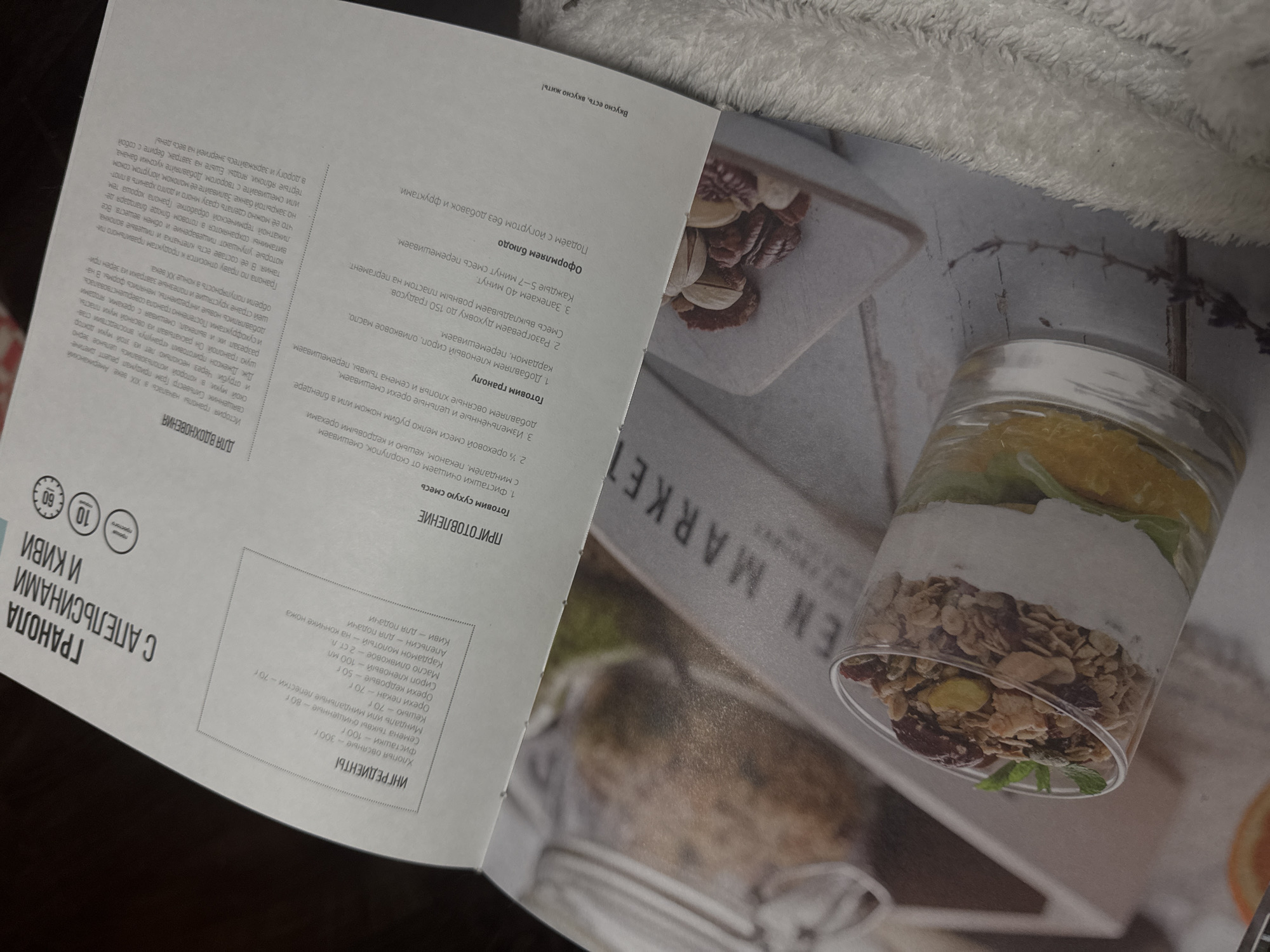 Here is a description of the image:
The image shows an open cookbook with a recipe for granola with oranges and kiwis. On the right page, there is a close-up photo of a clear glass filled with layers of granola, yogurt, and orange and kiwi slices. The granola appears to be made with oats, nuts, and dried fruit. The text on the left page contains the recipe ingredients and instructions in Russian. The book lies at an angle, and the background is blurred. It is impossible to pinpoint the location in Izhevsk, Russia, based on the information contained in the image.