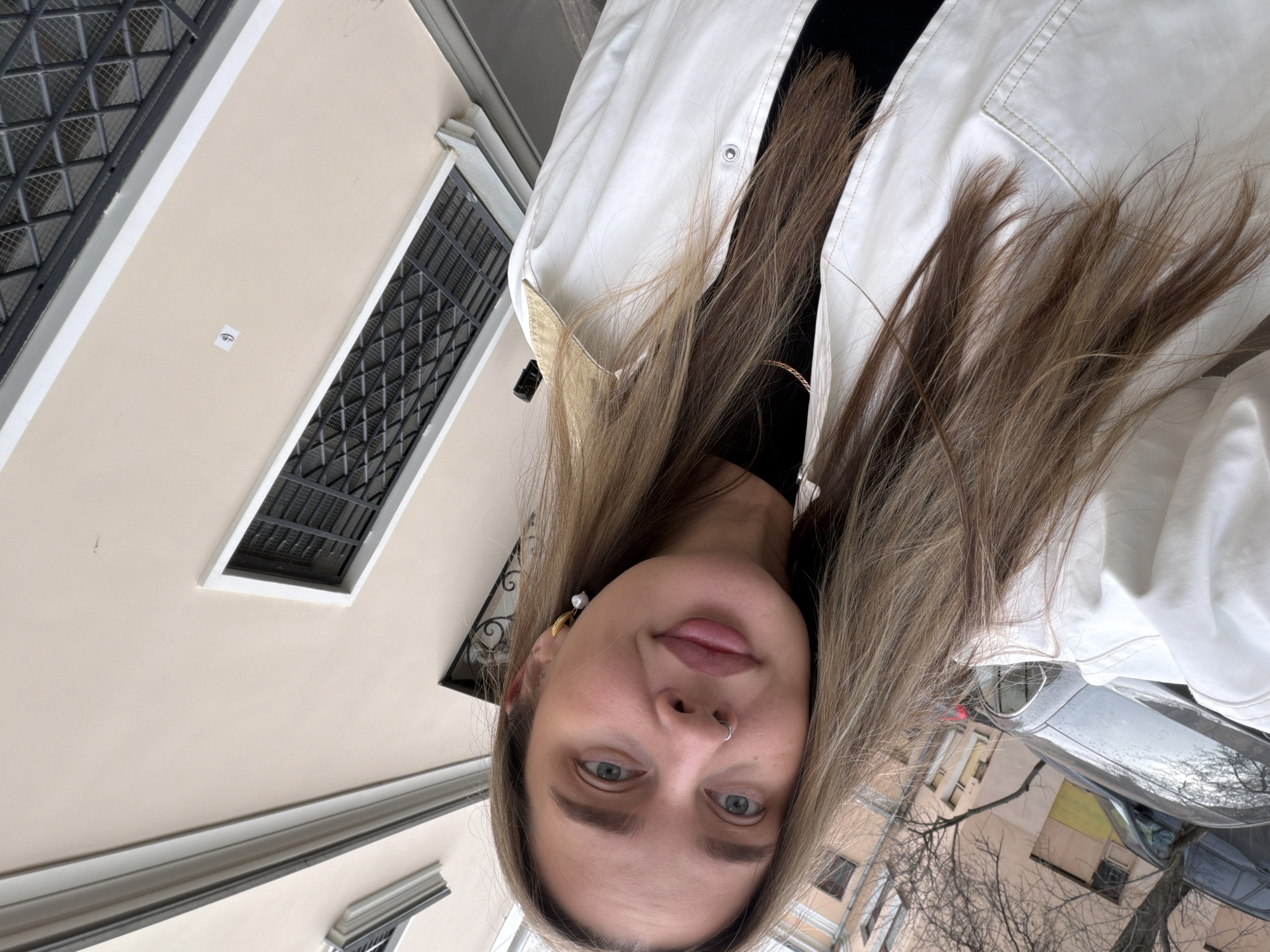 The image is a rotated selfie of a young woman taken outdoors in an urban environment. Her head is tilted, and she looks directly at the camera with a neutral expression. She has long, light brown hair with blonde highlights, which appears somewhat windswept. She wears a white jacket with visible stitching and a snap button, over a black top. Her accessories include a silver hoop nose piercing and a gold-toned earring with a white pearl on her left ear.
The background primarily features the exterior of a light beige or cream-colored building. The building has several rectangular windows covered by dark, ornate metal grates. A small, white sticker with a stylized black eye logo is visible on the building wall. In the lower background, a street scene is partially visible, including a silver car, bare tree branches, and other buildings, suggesting a cool, possibly overcast, daytime. The setting is an urban street in Moscow, Russia.
