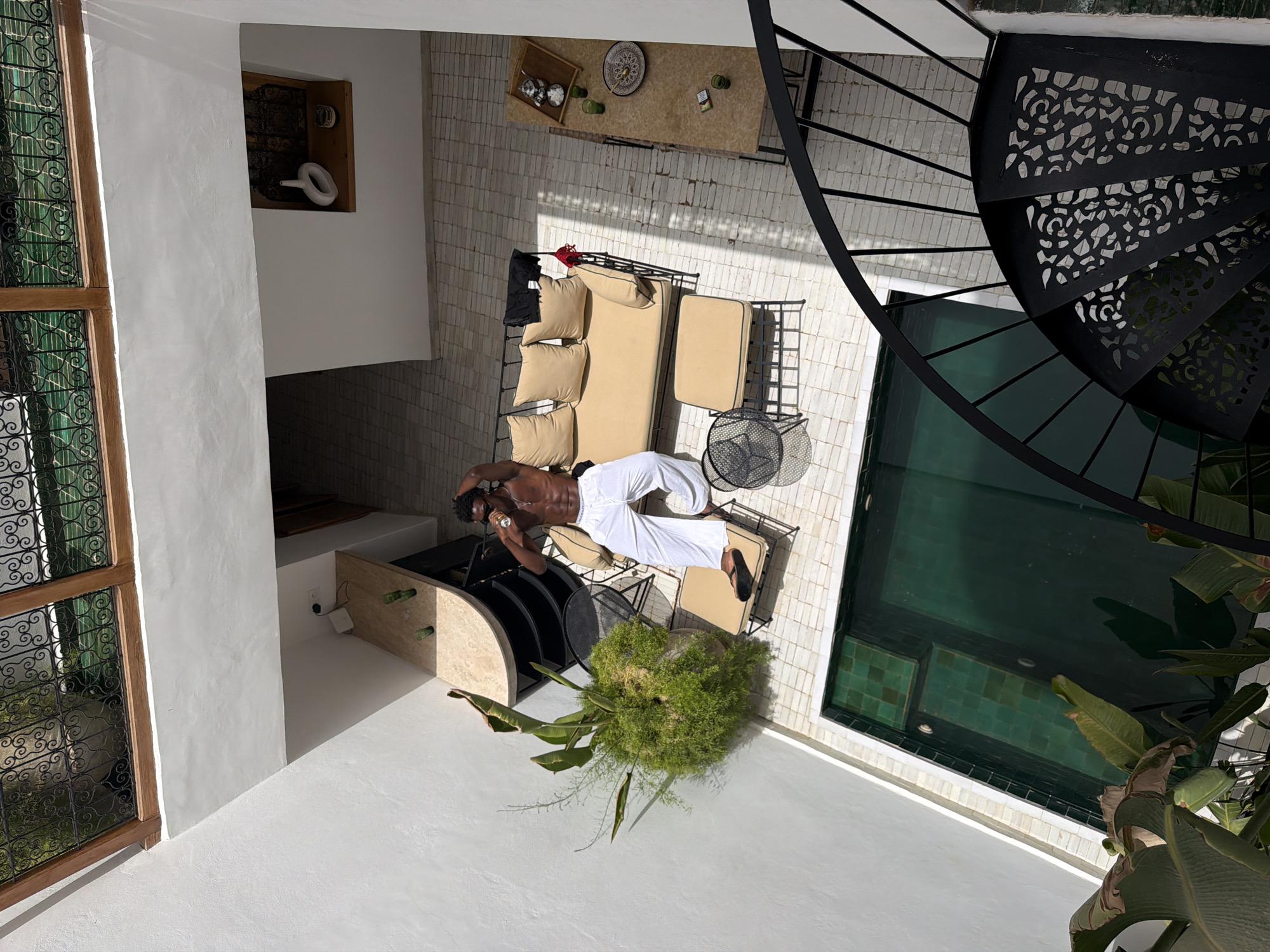 The image depicts a man in white pants and no shirt, holding a drink and relaxing on a tan outdoor sofa in a sunny, tiled courtyard. To the left of the sofa is a large potted plant. A small, dark pool with green tiles is situated in the foreground. In the background, there is a table with a teapot, cups, and other items, as well as a spiral staircase on the right. The architecture suggests a traditional Moroccan riad in Marrakech.