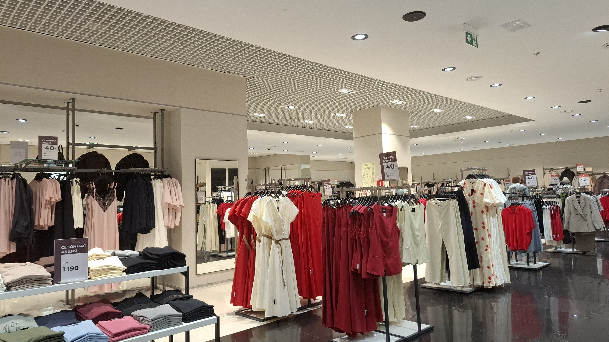 Here is a detailed description of the image:
**Location:** The image appears to be taken in a clothing store within a shopping mall. Based on the text visible on signage in the image, it is likely located in Saint Petersburg, Russia.
**Environment:** The store has a high, white ceiling with a grid-like structure. The lighting is bright and diffused. The floor is a glossy black, possibly polished stone or tile.
**Subjects and Objects:**
* Clothing racks: Multiple racks are filled with clothes, mostly dresses, blouses, and pants in various shades of red, white, and neutral colors.
* Signage: Several signs indicate sales and discounts, mentioning phrases such as "СЕЗОННАЯ АКЦИЯ" (seasonal sale) and "-40%". One sign also displays a price starting from 1190.
* Clothing displays: There is a table with folded shirts or sweaters in different colors.
* Mirror: A mirror reflects other clothing displays.
* Ceiling fixtures: Lighting fixtures and what appears to be an emergency exit sign are visible on the ceiling.
* People: A woman wearing a blue sweater is located deeper into the store.
**Actions and Interactions:** There are no immediate actions or interactions depicted in the image. The scene captures a static moment inside a clothing store, likely during shopping hours.
**Overall Impression:** The image showcases a well-lit, organized retail space offering a variety of women's clothing. The sales signage suggests a promotional period.