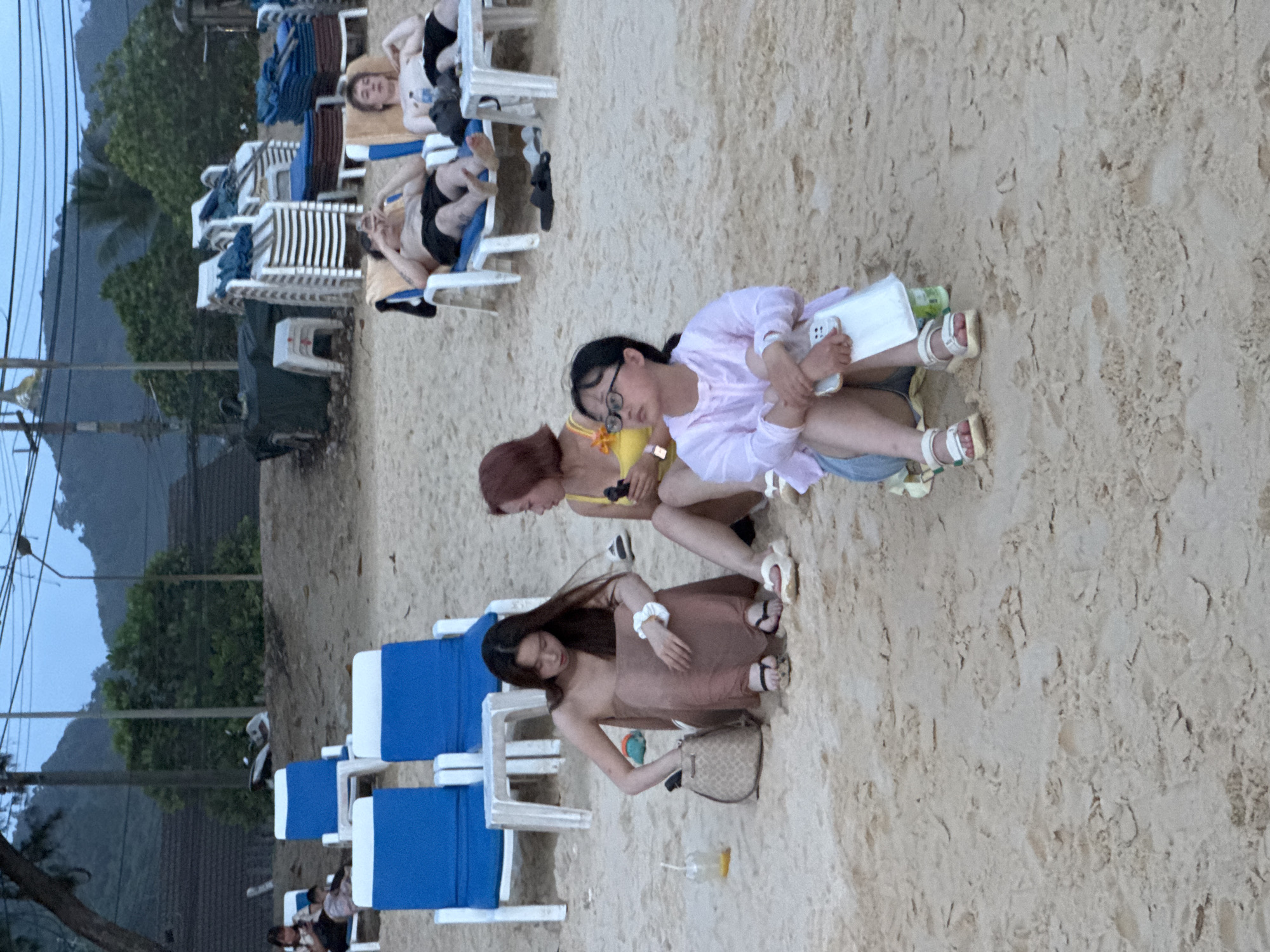 The image depicts a sandy beach scene in Karon, Thailand, likely during the late afternoon or early evening. In the foreground, three young women are seated on the sand. The woman on the far left, with long dark hair, is wearing a long, light brown dress and appears to be reaching into a patterned bag. Beside her is another woman with reddish-brown hair, wearing a yellow bikini top. The third woman, in the foreground right, is wearing glasses, a light pink sheer top, and denim shorts; she is holding a white object, possibly a phone, and looking to the left. Further back, two men are relaxing on lounge chairs. One man, with dark hair, is lying on his back and looking at his phone, while the other, with lighter hair, is propped up on his elbows, looking towards the left. Several blue and white lounge chairs are arranged on the beach, and stacks of chairs and beach equipment are visible in the background. The environment suggests a resort or public beach area, with trees and structures indicating a coastal town. The lighting suggests the sun is low, casting a diffused light over the scene.