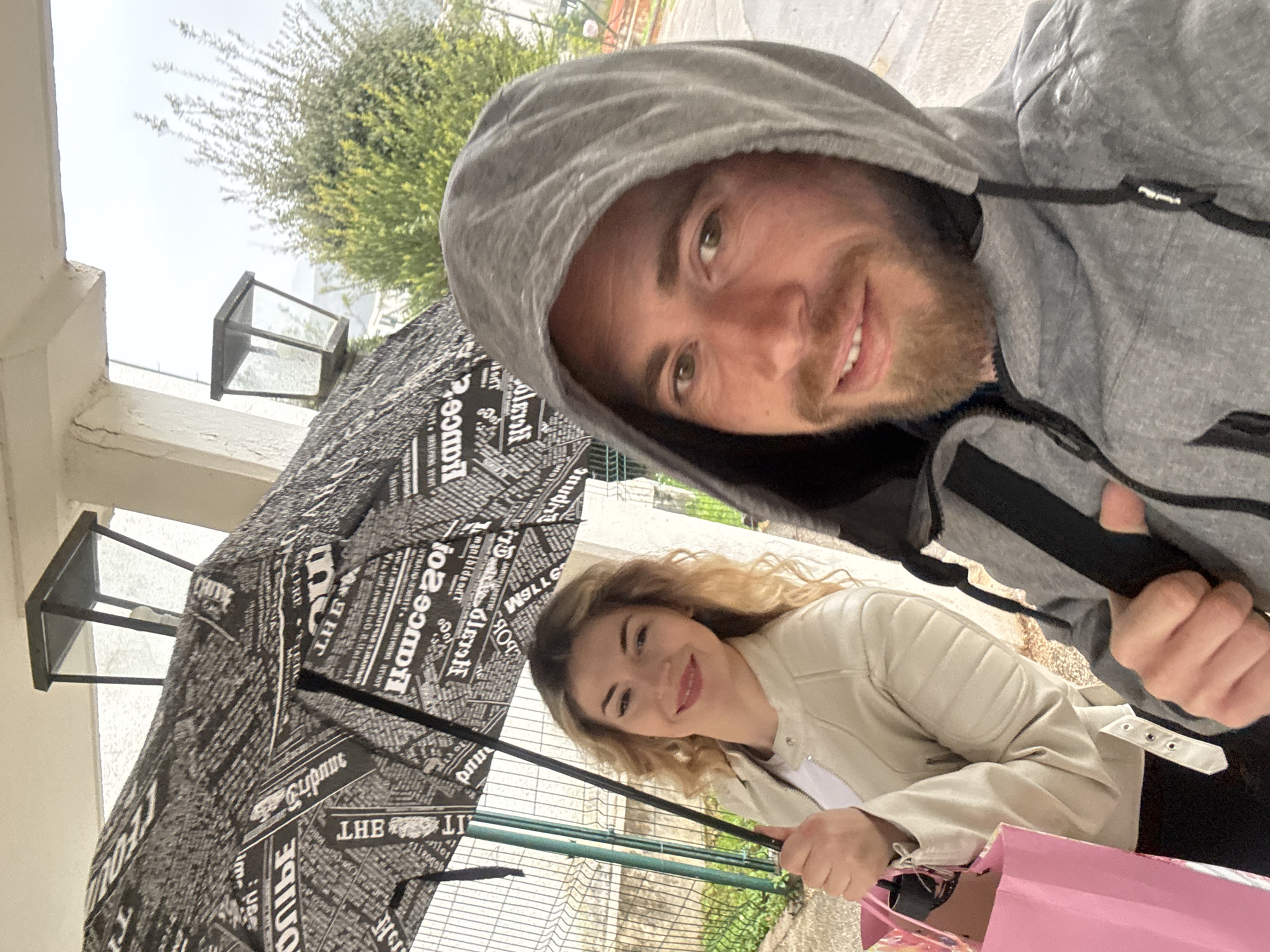 The image depicts a selfie of a man and a woman under an umbrella covered in a newspaper-print design, likely taken outdoors on an overcast day. The man is wearing a grey hooded jacket and smiling into the camera. The woman is smiling, holding the umbrella, wearing a light-colored jacket, and a pink shopping bag is visible. In the background, there is a building structure, greenery, and a fence. The location is suggested to be Budva, Montenegro, but this is not explicitly visible in the image.