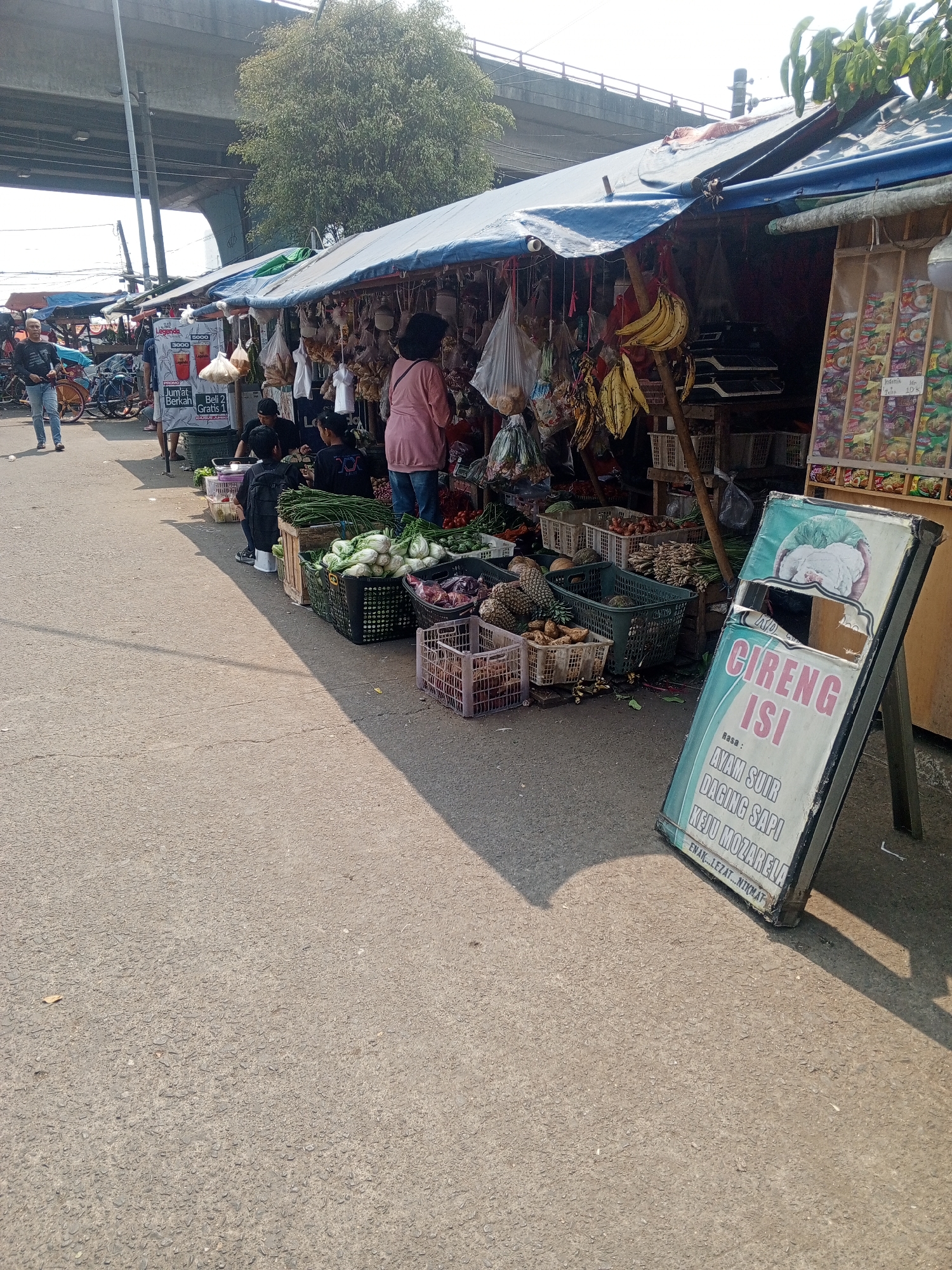 A busy street-side market in Depok, Indonesia, is captured under an overpass. Stalls are set up along the asphalt road, displaying a variety of goods. In the foreground, a weathered A-frame sign advertising "CIRENG ISI" (filled fritters) with flavors like chicken, beef, and mozzarella is propped up. Beyond the sign, produce such as cucumbers, leafy greens, pineapples, and onions are neatly arranged in baskets. Several individuals are seen interacting around the stalls, including a woman in a pink top and jeans looking at goods, and others seated or standing amidst the merchandise. Some bicycles are parked in the background, indicating pedestrian and vehicle traffic. The environment is characterized by the bright sunlight casting long shadows, suggesting it is daytime. The overall scene depicts a vibrant local marketplace with a mix of commercial activity and everyday life.