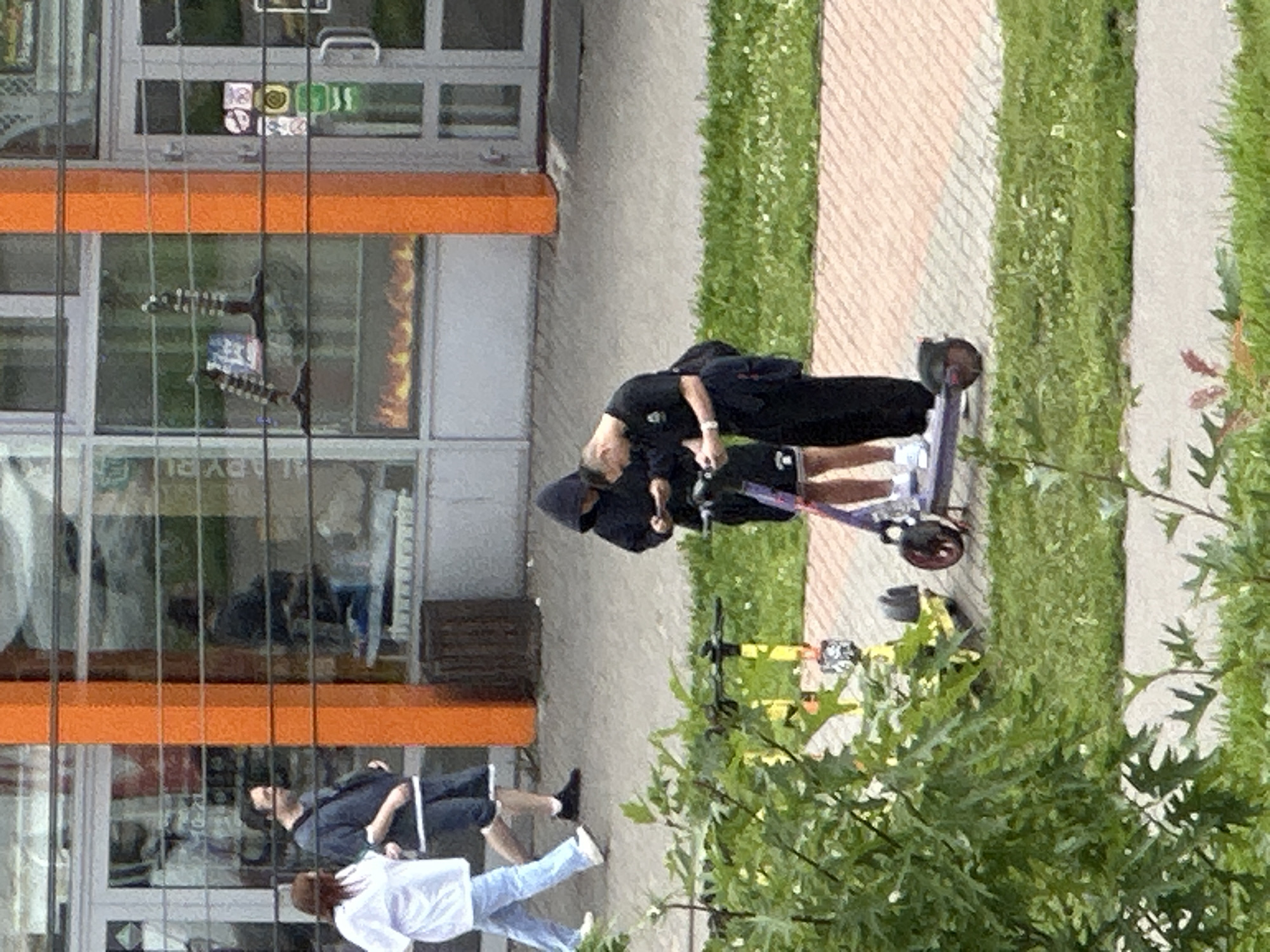 Two young men, likely in their late teens or early twenties, are standing on a paved walkway outside a building with an orange facade. One man is wearing a black hooded sweatshirt and the other is in a black t-shirt and pants. Both are looking down at their cell phones, and appear to be interacting with them. They are positioned near two parked electric scooters, one yellow and one purple, which are on the walkway. In the background, several people are walking on the sidewalk in front of a storefront. The environment appears to be a commercial area in the city of Podolsk, Russia.