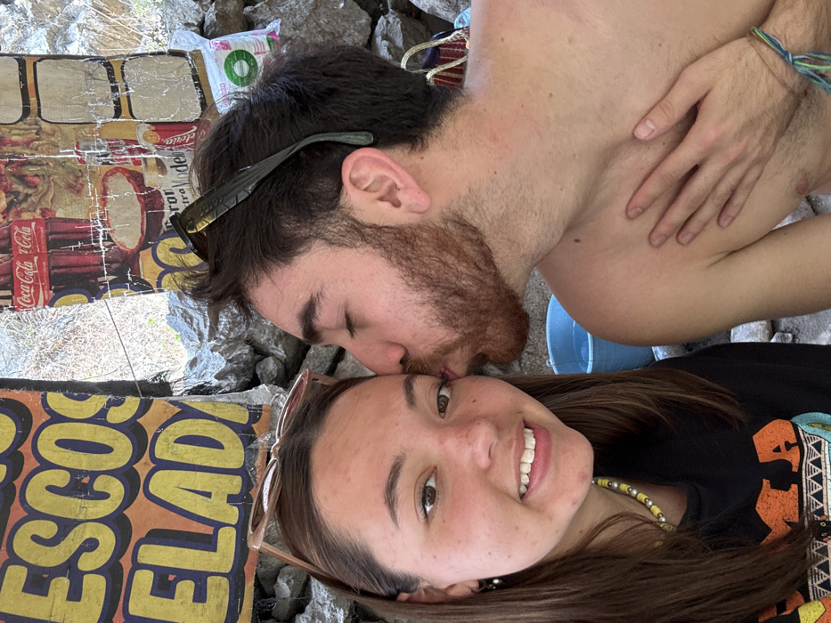A close-up image depicts two young adults in an outdoor setting in San Miguel Atlapulco, Mexico. On the left, a female with medium-brown hair, light brown eyes, and a light complexion smiles directly at the camera. She wears a black top with an orange and yellow design, a yellow beaded necklace, and small silver hoop earrings. Light-colored sunglasses are perched on her head. To her right, a male with dark hair and a reddish-brown beard and stubble is shown with his eyes closed, kissing the female's left cheek. He appears shirtless, with dark sunglasses resting on his head, and a multi-colored braided bracelet visible on his left wrist. His left hand rests flat against his upper chest. The background features a textured stone wall or rock formation. Several weathered commercial signs and posters are affixed to the wall, including a yellow sign with blue lettering partially legible as "ESCOS ELADA" and other posters featuring "Coca-Cola" and "Modelo" branding. A light blue bucket, partially filled with water, is visible in the mid-ground behind the male's arm, and a white bag is visible in the upper right background.