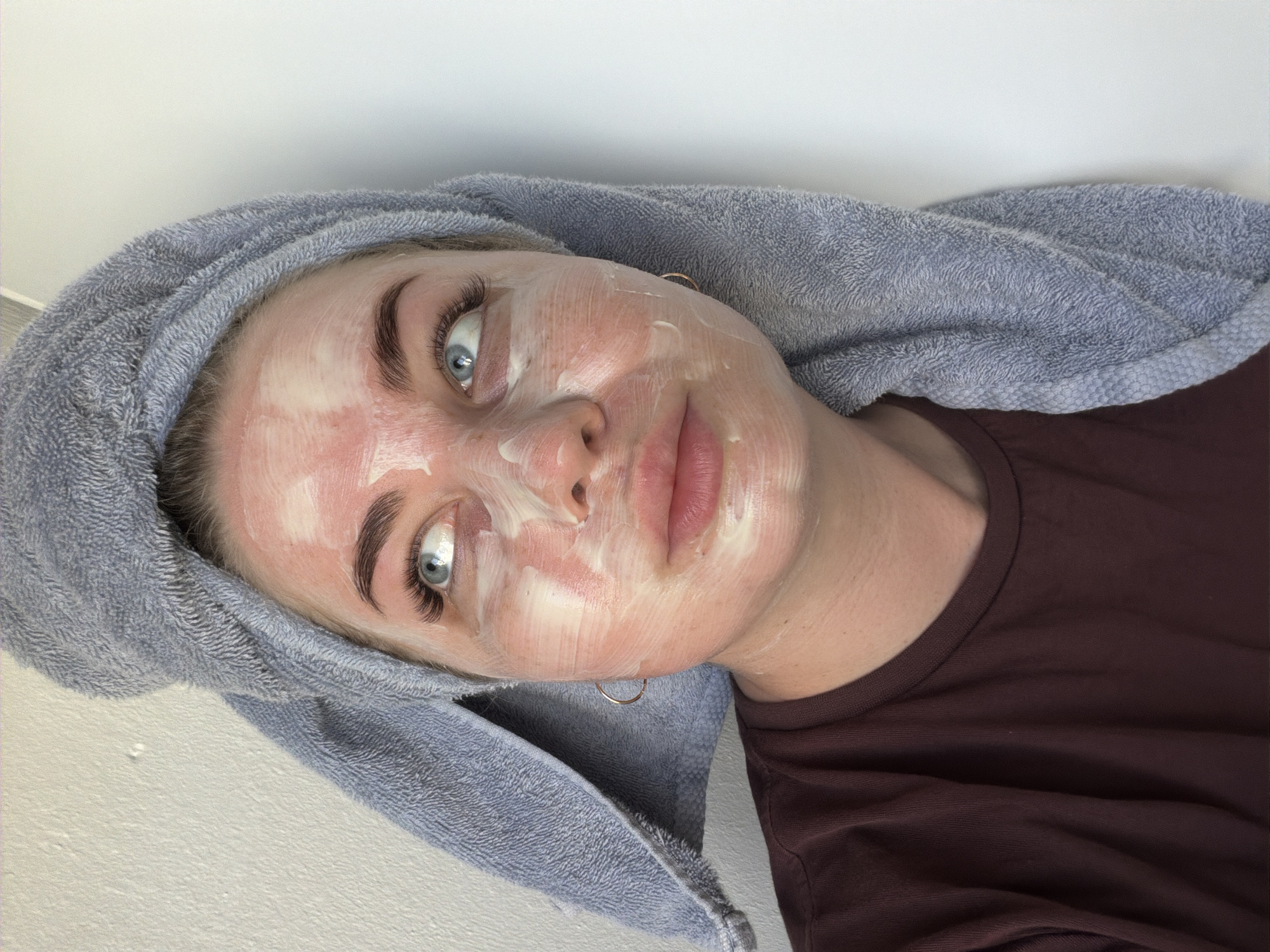 A woman with blue eyes and light skin, wearing a brown shirt and a gray towel wrapped around her head, is lying down. Her face is covered in a white substance, possibly a facial mask. She has gold hoop earrings. The background is a plain white wall. The location is not discernible from the image.