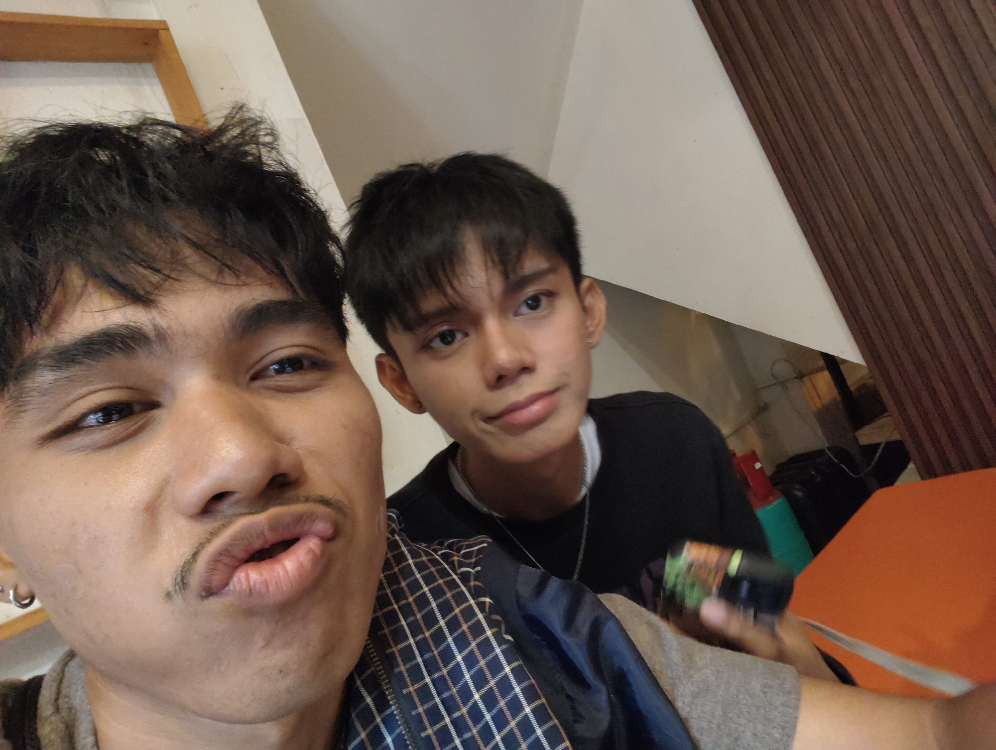 The image is a selfie taken by two young men in what appears to be a cafe or restaurant. The person in the foreground is making a kissy face, with their mouth pursed and a thin mustache visible above their upper lip. They are wearing a grey hoodie and a checkered jacket. To the left, a silver hoop earring is visible. The second man, in the background, is wearing a black t-shirt and a silver chain necklace. He is looking towards the camera with a neutral expression. In the background, there are wooden panels, a white wall, and an orange table. The lighting suggests an indoor setting, possibly during the day. The presence of a small, dark-colored object, potentially a container, held in the foreground individual's hand is noticeable. The overall atmosphere is casual and friendly. Based on the context of similar images and common tourist spots, it's plausible this location could be within a cafe in areas like Poblacion, Davao Oriental, known for its burgeoning food scene, or perhaps a cafe in the city center like those found near Roxas Avenue, Davao City.