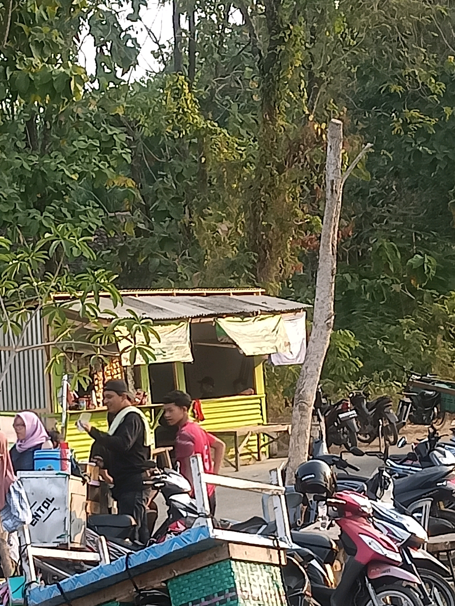 The image depicts a street scene in Sambiduwur, Indonesia, with a small roadside stall and several motorcycles. In the foreground, a blue and white cooler with the text "ENTOL" is visible, along with stacks of wood and a green woven mat. To the right, a red motorcycle is parked. Behind the stall, a few motorcycles are lined up.
Three individuals are present. A woman wearing a pink headscarf stands on the left, next to the cooler. In the center, a man in a black long-sleeved shirt and a black cap is holding what appears to be a piece of paper. To the right of him, a young man in a red shirt is looking towards the viewer.
The stall itself has a green facade with a corrugated metal roof covered by a white tarpaulin. The background is dominated by lush green trees and foliage, suggesting a semi-rural or tropical environment. A dry, weathered tree trunk stands prominently in the middle ground. The overall impression is a casual, everyday scene at a local gathering spot.