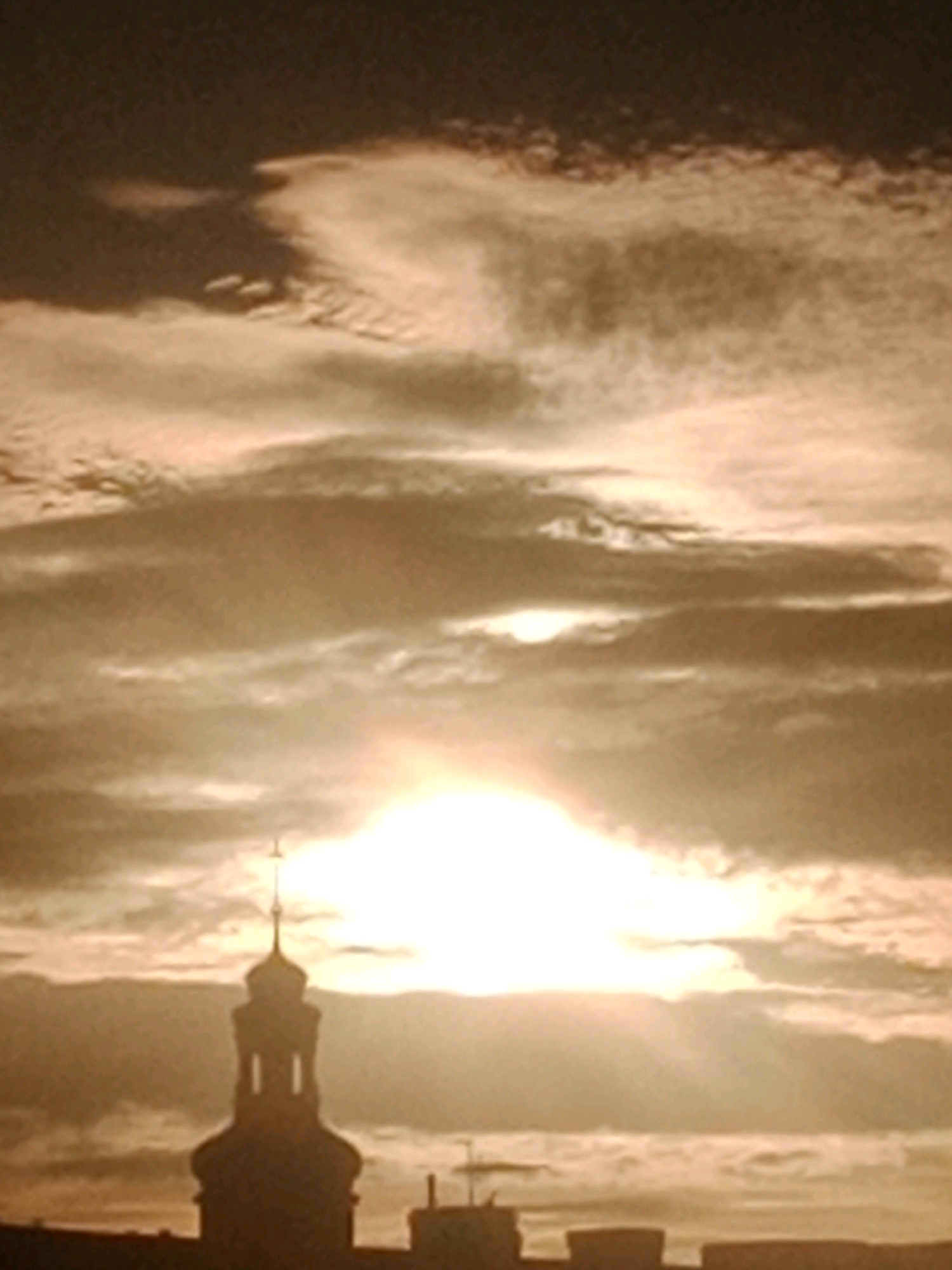 The image depicts a silhouetted church steeple rising above buildings against a backdrop of dramatic, sun-drenched clouds. The sun, appearing as a bright, diffused orb, is partially obscured by the clouds, casting a warm, golden light across the sky. The sky itself is filled with textured, wispy clouds, creating a sense of depth and movement. The architecture of the steeple, with its rounded dome and tall spire, suggests a historical building. The overall impression is one of a serene and majestic urban landscape at dawn or dusk, possibly in a European city like Bydgoszcz, Poland, known for its historic churches and characteristic skyline.