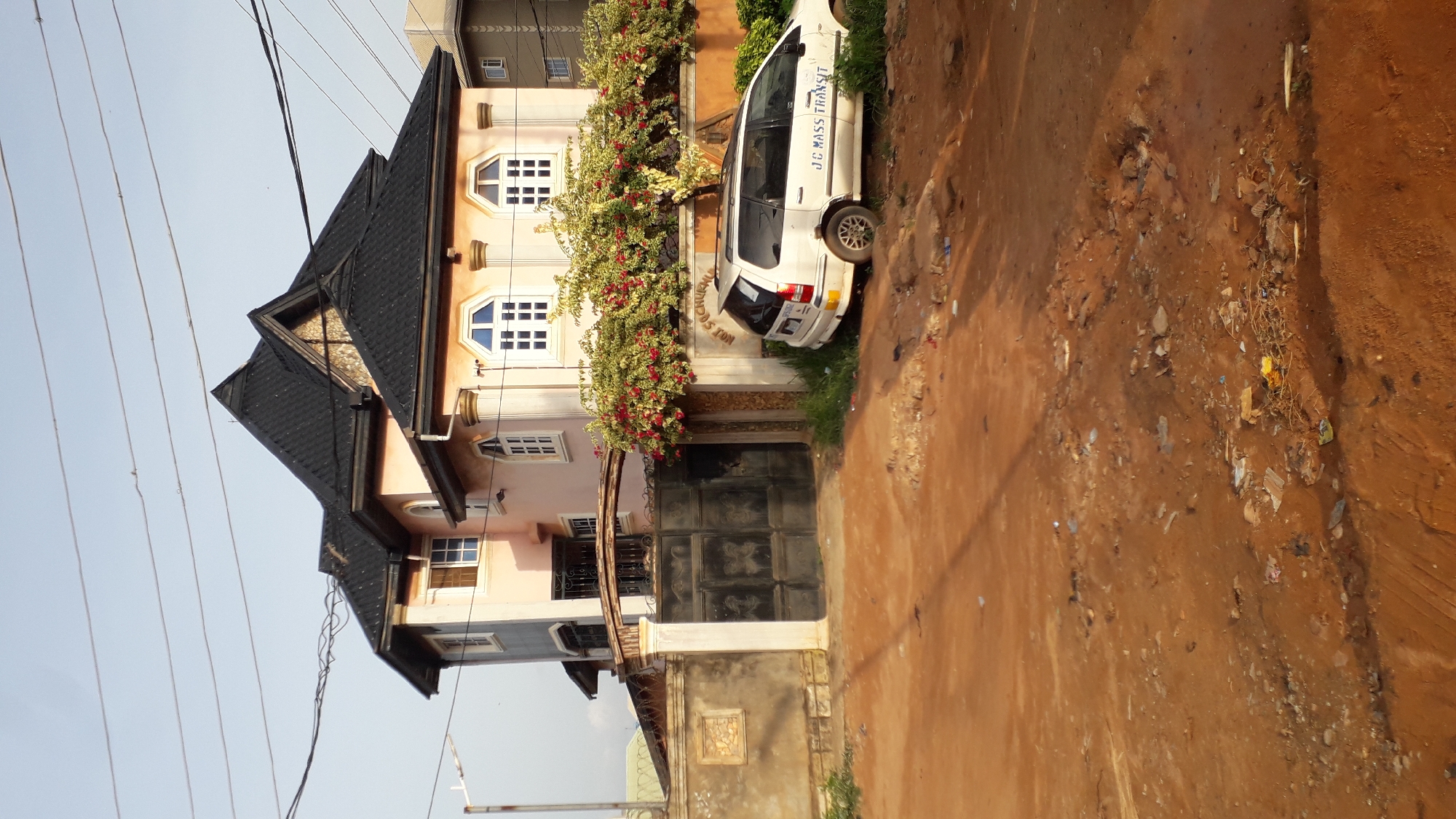 The image depicts a multi-story residential building in Nkpor, Nigeria. The house is painted in shades of light pink and blue, with a black shingled roof. The facade features white framed windows. A dark metal gate secures the entrance, adorned with flowering vines on top. A white minivan with "JE MASS TRANSIT" printed on its side is parked next to the gate, partially obstructing the view of the building. Overhead power lines are visible against a pale blue sky. The ground in front of the building is bare, dry, and littered with debris.