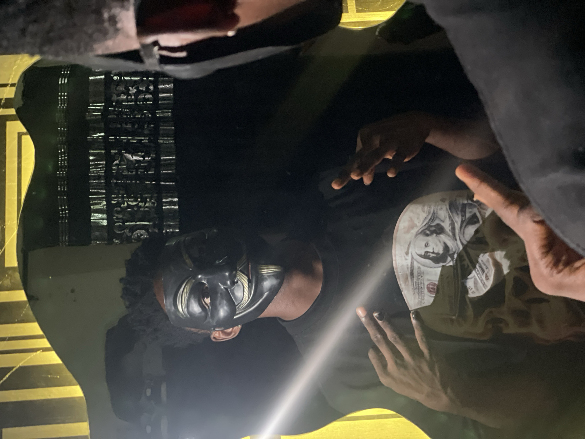 The image depicts a reflection of a person wearing a black mask, likely inside a vehicle, with money visible. Lokoja, Nigeria, is not discernible from the visual information provided. The environment is dimly lit, possibly nighttime. The reflection is on a glossy surface, potentially a table or car dashboard, which also shows the reflection of currency, possibly United States dollar bills. An individual's hand is visible near the currency.