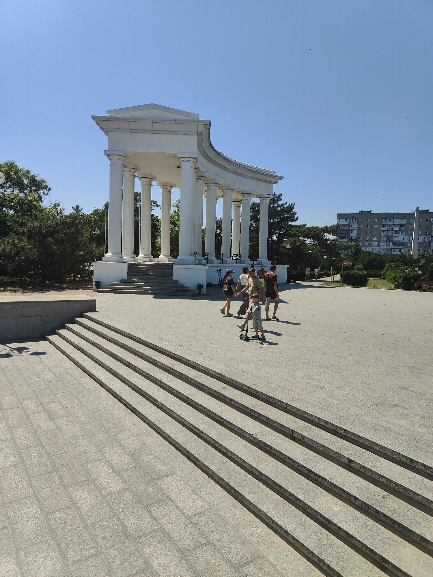 The image shows a sunny day in a public space in Chornomorsk, Ukraine, featuring a large, white, neoclassical rotunda with multiple columns. In the foreground, a wide set of stone steps leads up towards the rotunda, with a paved area to the right. A young boy is riding a scooter on the paved area, and several adults are walking in a group near the rotunda. Behind the rotunda, there are trees and a residential building. The sky is clear blue with a few distant birds.