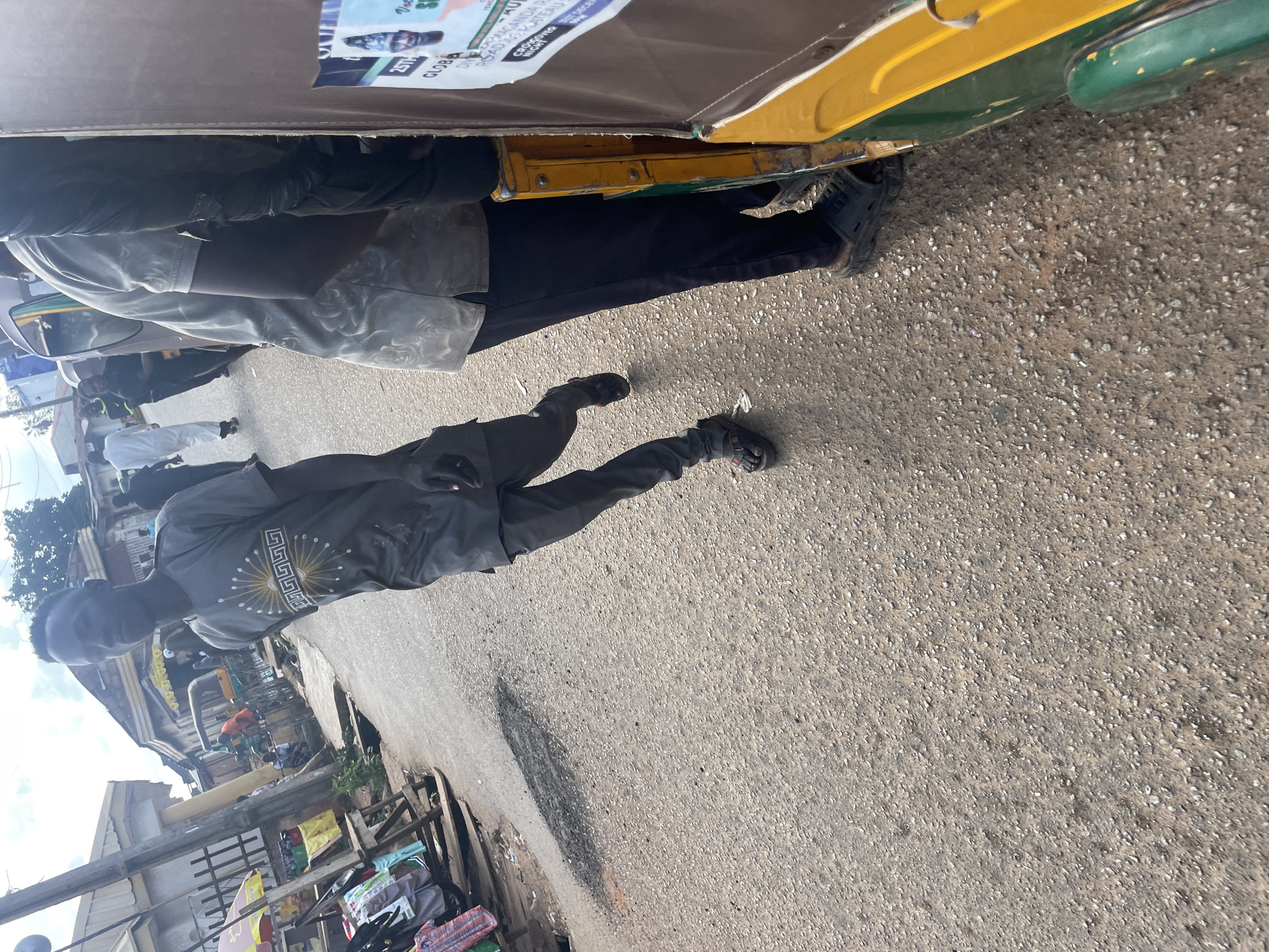 In Gura Top, Nigeria, a young man in a grey t-shirt with a sunburst design and dark pants walks down a paved road. He is looking towards the camera, and his right hand is extended slightly. To his left, a rickshaw-like vehicle is partially visible, with its green and yellow body a prominent feature. In the background, a market scene unfolds with stalls and other individuals, some walking and others gathered. The sky is overcast, with clouds dominating the upper portion of the image. The road surface is rough, indicating a well-worn urban environment.