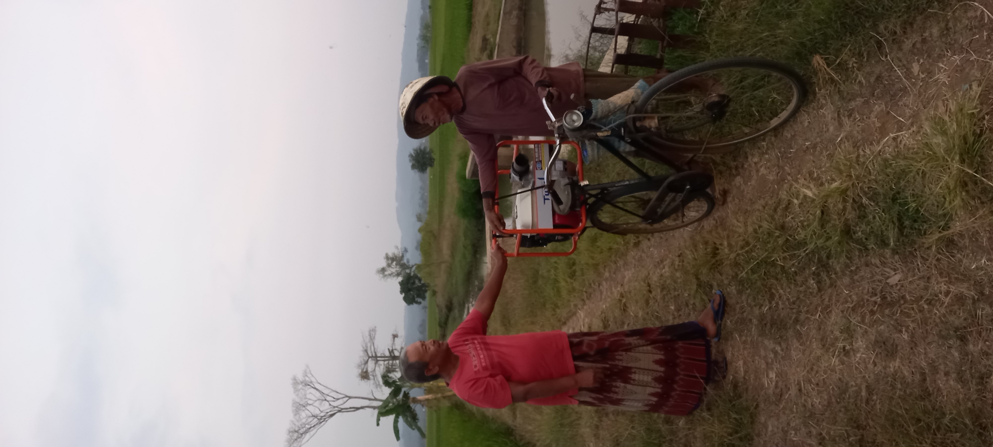 Two elderly Indonesian men are standing on a dirt path in a rural area, likely Sidamulya, Indonesia, with rice paddies and hills in the background under a hazy sky. The man on the left, wearing a red t-shirt and a patterned sarong, is facing right, looking at the other man. The man on the right, wearing a brown long-sleeved shirt and a conical hat, is facing left, looking at the first man. Between them is a black bicycle with a large orange rack on the back carrying a white and silver portable pump or generator. The man on the left has his right hand extended towards the bicycle, while the man on the right has his left hand resting on the bicycle. The environment is grassy with some dry patches of dirt.