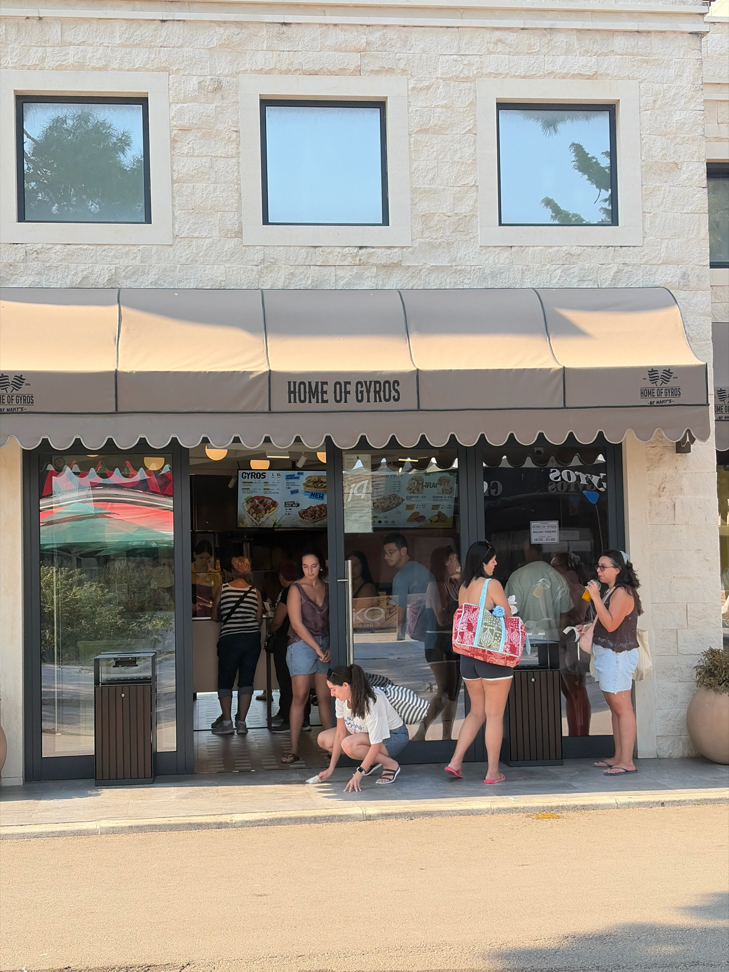 This image captures the exterior of a "Home of Gyros" restaurant in Budva, Montenegro. The establishment features a light-colored brick facade with three rectangular windows above a prominent beige awning that displays the restaurant's name. Underneath the awning, a glass-fronted storefront reveals the interior, where a digital menu board showcases various gyros options. Several people are visible in and around the entrance. Some are queuing inside, while others stand outside. A woman in the foreground is crouched down, appearing to clean or pick something up from the pavement. Another woman to her right holds a drink and a bag. The ground in front of the restaurant is paved, with a street visible in the lower portion of the frame. A brown trash receptacle is situated to the left of the entrance. The overall scene depicts a sunny day with clear skies reflected in the windows.