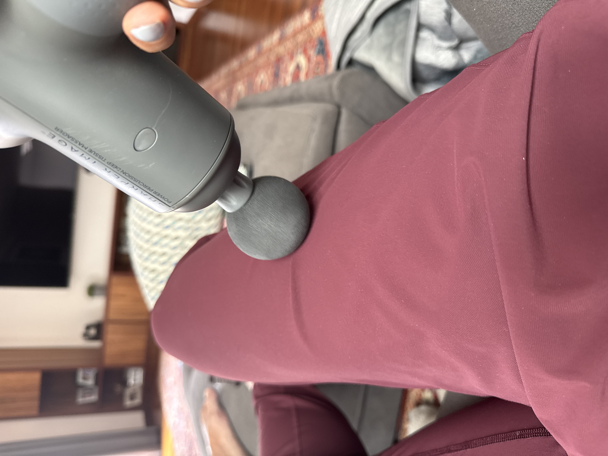 A person is using a Sharper Image power percussion deep tissue massager on their leg. The person is wearing maroon athletic pants. The background includes a rug, a grey couch, and a TV cabinet. The location is not in San Bernardino Tlaxcalancingo, Mexico.