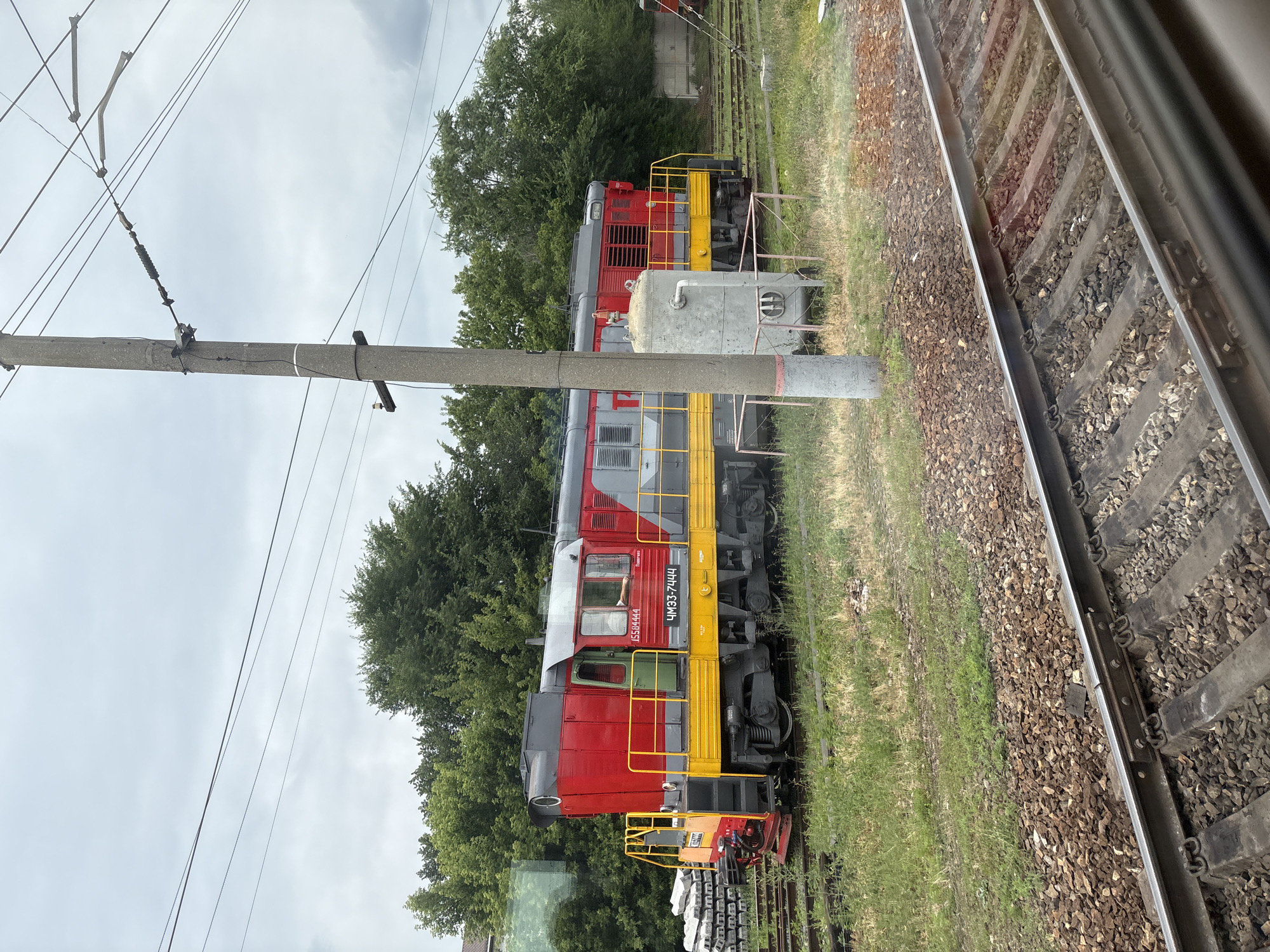 The image shows a red and gray diesel locomotive with yellow accents parked on a railway track. The locomotive has Cyrillic lettering on its side, including "ЧМЭ3-7444". A person can be seen through the locomotive's window, with their arm resting on the sill. In the background, there are trees, a utility pole with overhead wires, and a large cylindrical metal tank. The scene appears to be in a railyard or industrial area, with additional railway tracks and ballast visible in the foreground. The sky is overcast. The provided text in the image is in Russian. The location of Povorino, Russia, is mentioned in the prompt.