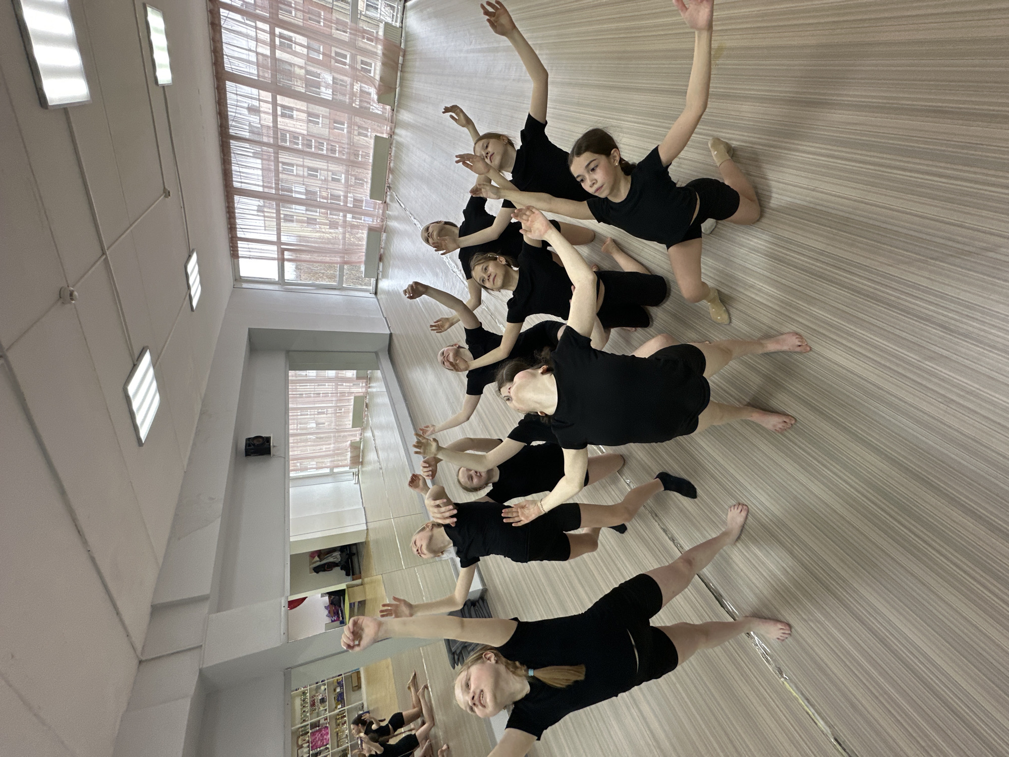 **Location:** A dance studio in Votkinsk, Russia.
**People:** Eight young female dancers are practicing a routine.
**Subjects:** The dancers are dressed in black leotards and shorts. Most are barefoot, while one wears beige dance shoes and another black socks.
**Objects:** The studio has a light-colored wood-look floor, white walls, and a mirrored wall on one side. There are large windows with a view of buildings outside. The ceiling has fluorescent lights.
**Actions:** The dancers are posed in a line, with one arm raised and the other extended to the side. Their positions vary slightly, indicating different stages of a movement.
**Interactions:** The dancers appear to be working together, following the same choreography.
**Environment:** The dance studio is a bright, open space, typical for rehearsals and dance classes.