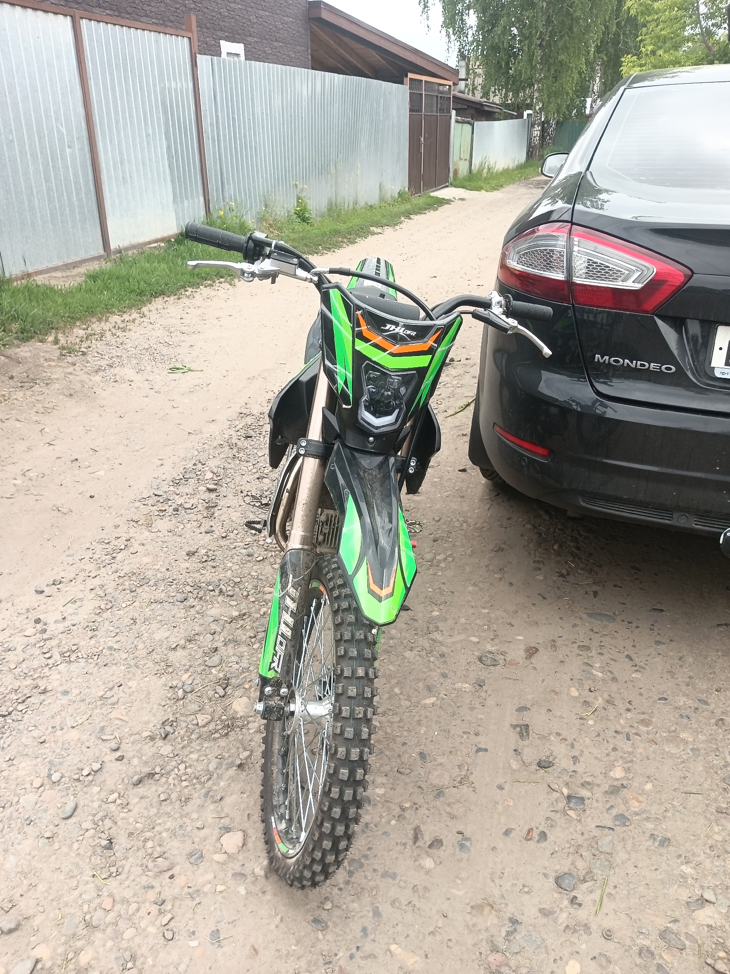 In an outdoor scene, a green and black dirt bike is parked on a gravel and dirt surface. The dirt bike is positioned in the foreground, facing forward, with its front wheel prominent. To the right of the dirt bike, the rear of a black Ford Mondeo sedan is visible. The car has its taillights illuminated and the "MONDEO" badge is clearly displayed. A metal fence runs along the left side of the image, behind the dirt bike. The background shows a building with a corrugated metal wall and a section of wooden fence. Trees are visible above the building in the distance. The ground is composed of dirt and small stones.