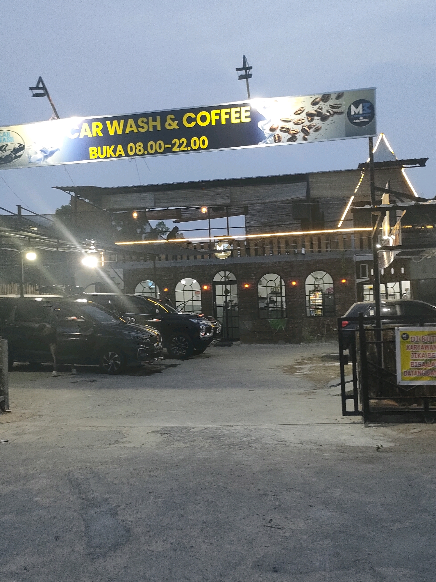 A sign above a car wash and coffee shop reads "CAR WASH & COFFEE BUKA 08.00-22.00". The shop appears to be located in Batam, Indonesia, judging by the context of the sign. Several dark-colored cars are parked outside the establishment, which has an open-air design with a covered roof supported by pillars. The building features arched windows and is illuminated by string lights. A person is visible standing near one of the cars, and another individual appears to be on the roof structure. A yellow sign with Indonesian text is mounted on a gate to the right of the image, likely providing further information about the business. The overall scene suggests an evening setting, possibly dusk, due to the lighting conditions.
