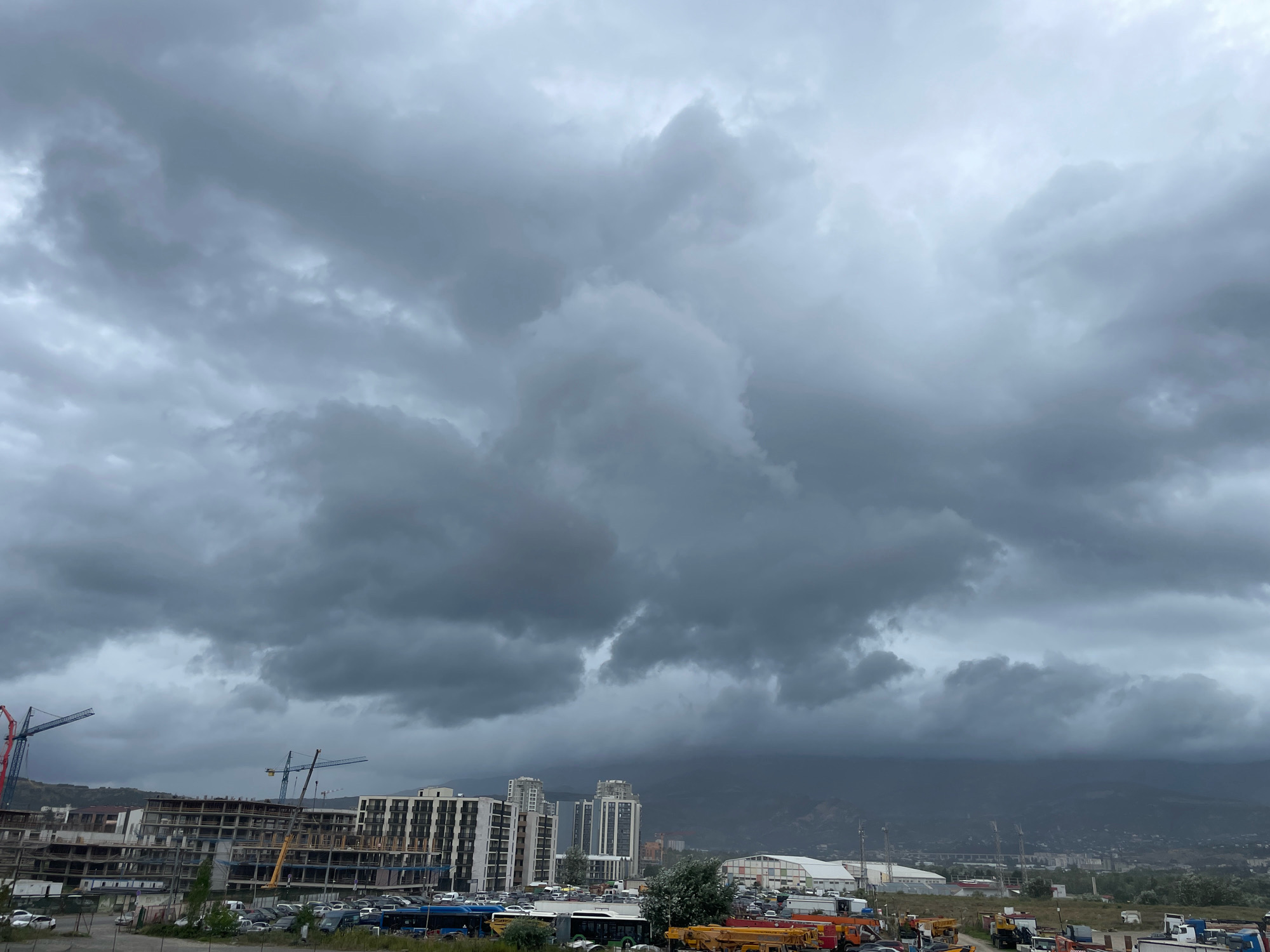 The image depicts a cloudy, overcast sky over a city landscape. In the foreground, construction sites are visible with cranes and unfinished buildings. Several cars and buses are parked in lots or driving on roads. In the background, a range of mountains can be seen through the hazy atmosphere. The scene appears to be in an urban area of Tbilisi, Georgia, likely a district undergoing development. The weather is somber, with heavy, dark clouds dominating the sky, suggesting a possibility of rain. No people are clearly visible in the image.