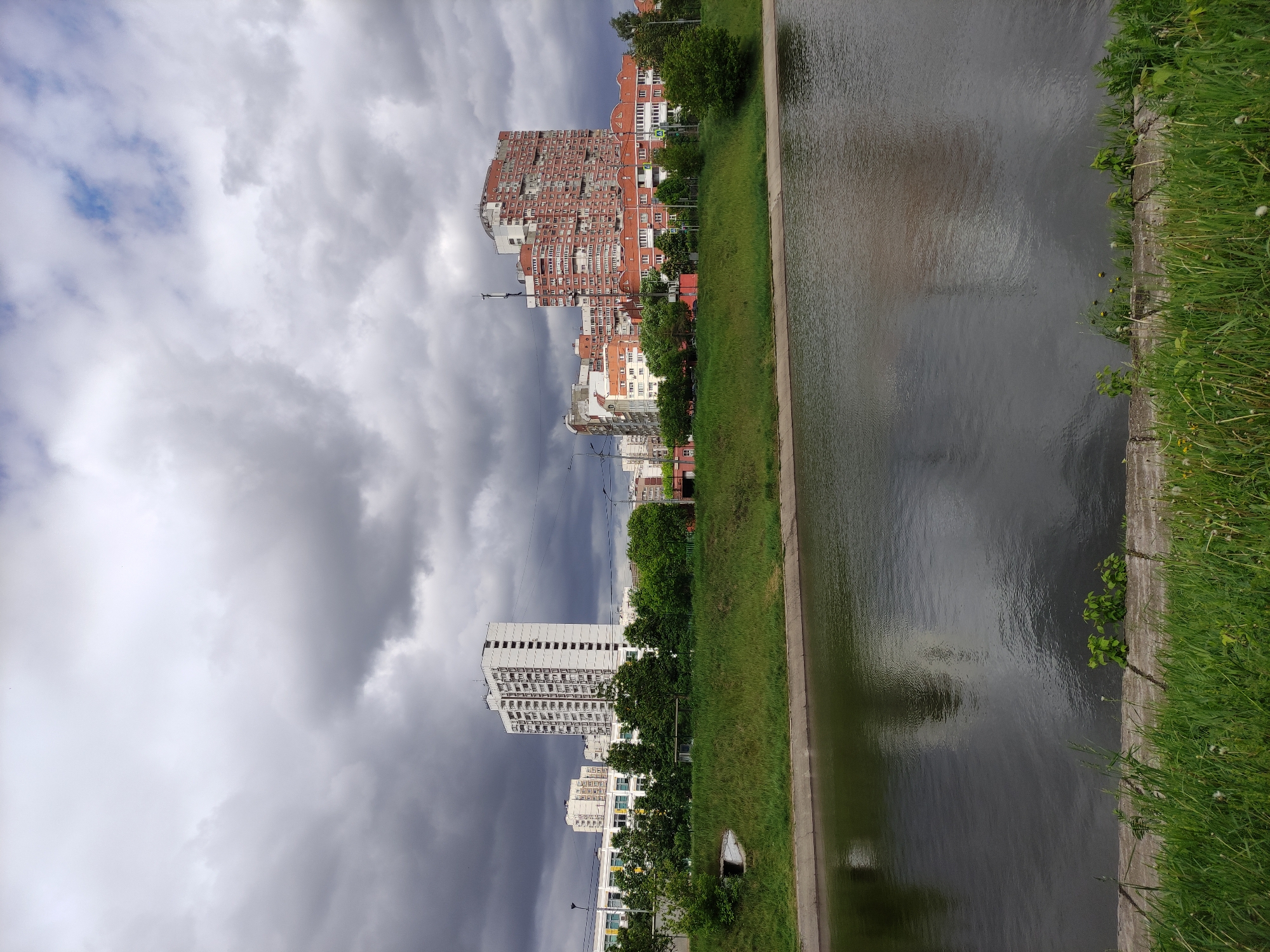 The image shows a cityscape alongside a body of water. The sky is filled with heavy, grey clouds, suggesting an overcast or stormy day.
On the left side of the image, several buildings of varying heights and designs can be seen. A particularly distinctive structure is a large, red-brick building with a unique, complex facade. Further down, there's a taller, white building with a more modern, rectangular shape. Trees line the street in front of these buildings.
To the right, a concrete canal runs parallel to the buildings. The water is dark, reflecting the cloudy sky. The edge of the canal is lined with green grass and vegetation.