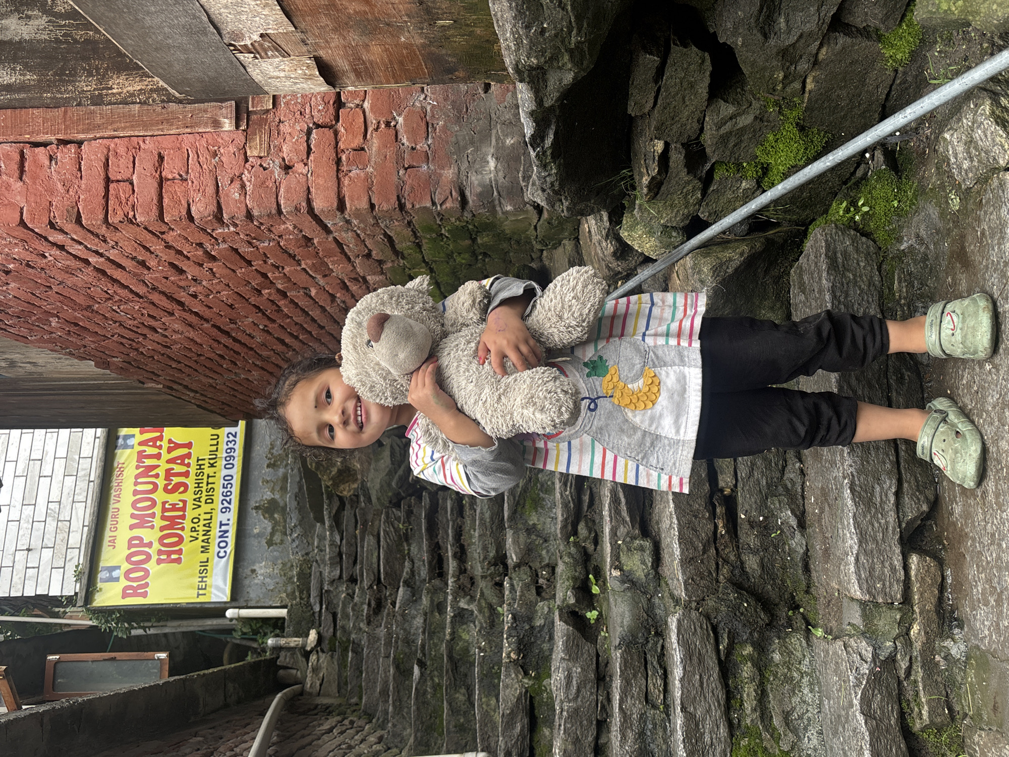 A young girl is standing on a stone staircase in Vashisht, India, holding a stuffed toy. She is wearing a grey long-sleeved shirt with colorful stripes and a picture of a pineapple on it, black pants, and green clogs. To her left, a yellow banner reads "ROOP MOUNTAIN HOME STAY" and provides contact information. The staircase is flanked by stone walls and some greenery. The overall scene appears to be an outdoor setting in a village or town.