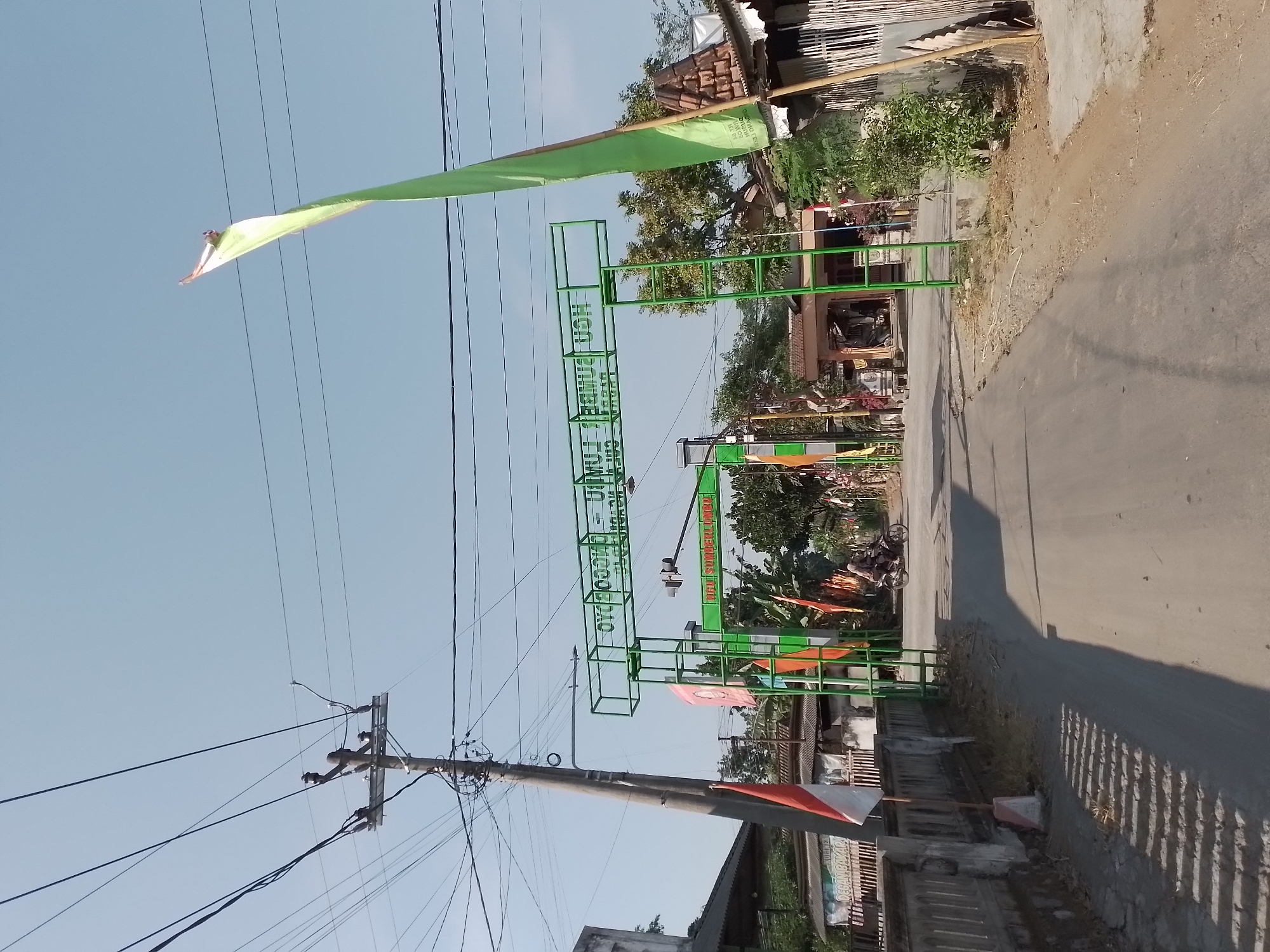 The image depicts a street scene in Ngancar, Indonesia. A large green archway spans across the road, bearing text in Indonesian which translates to "Welcome to Ngancar - Sumber Lumajang". On the left side of the road, a utility pole with numerous wires is visible, along with a concrete barrier and a building. The right side of the road features trees, a building with a thatched roof, and a tall green flag. A motorcycle with two riders is moving away from the camera down the road. The sky is clear and blue. The overall scene suggests a welcoming entrance to a local area or village, possibly decorated for an event.