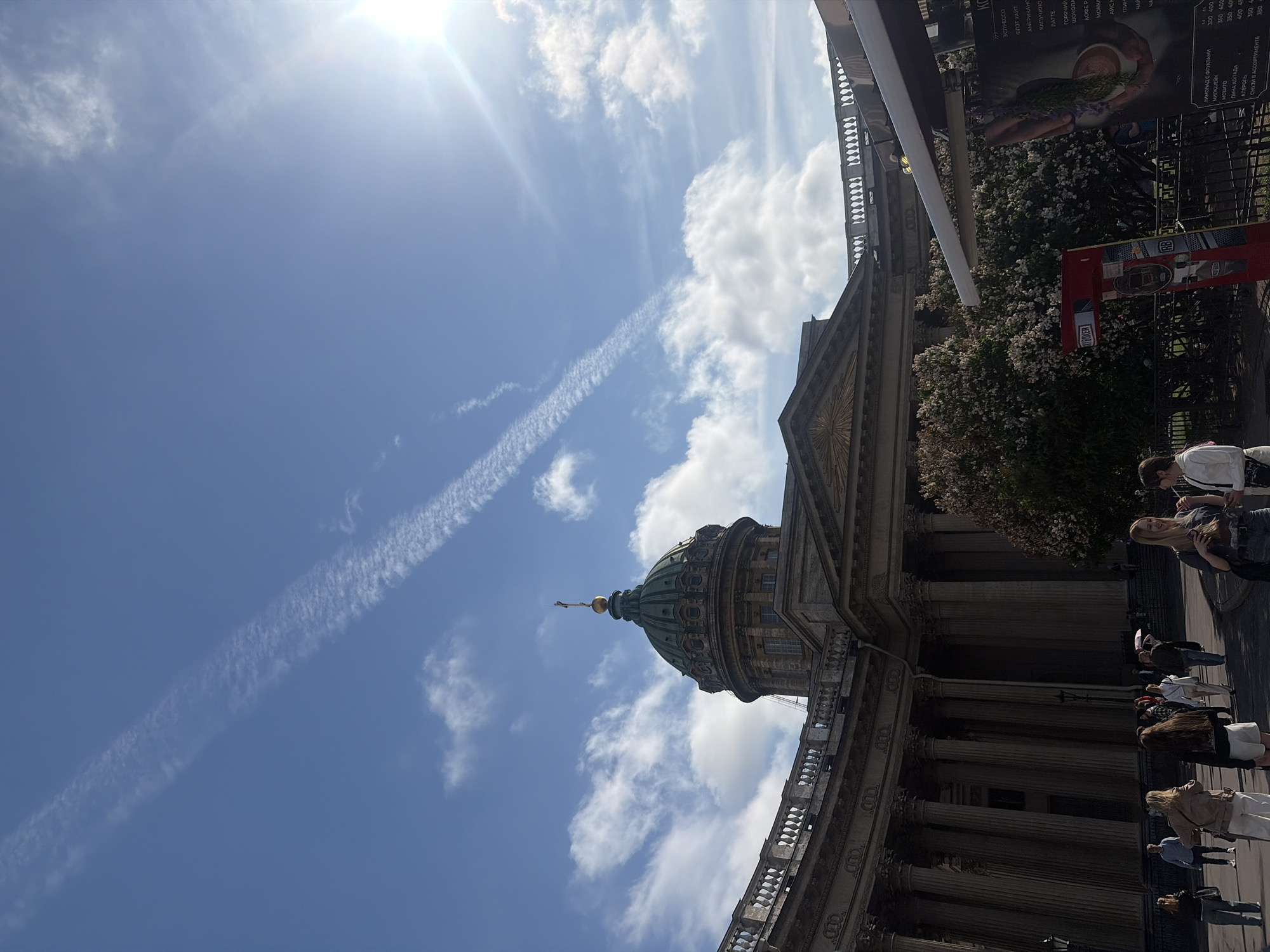The image shows the Kazan Cathedral in Saint Petersburg, Russia, on a sunny day with a clear blue sky and scattered clouds. The grand neoclassical cathedral, with its iconic colonnade and dome, dominates the background. In the foreground, a street scene unfolds with people walking, some appearing to be tourists. Several trees, some with white blossoms, are visible. A food stall or kiosk with a menu board is also present on the right side of the image. The overall atmosphere is bright and bustling, suggesting a daytime activity in a popular urban location.