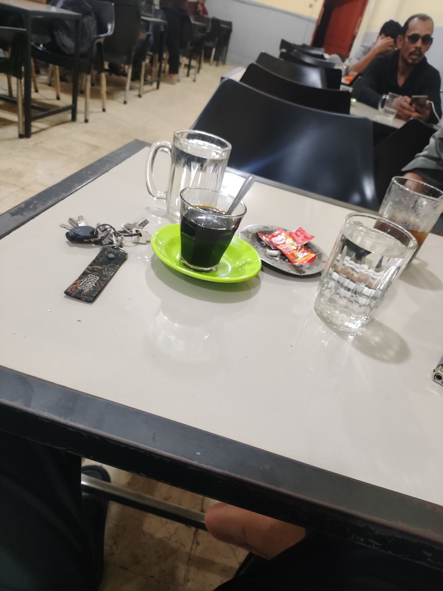 The image presents a candid view of a casual indoor setting, likely a cafe or eatery, situated in Lhokseumawe, Indonesia. The foreground is dominated by a light-colored table, displaying several items.
On the left, a set of keys is placed, attached to a black keychain with a decorative fob. The word "DEADPOOL" is clearly visible on this fob. Adjacent to the keys is a small glass cup containing a dark beverage, presumed to be coffee, resting on a vibrant green saucer. Behind this, a larger clear glass mug, filled with water, stands.
To the right, a small metallic ashtray holds several cigarette butts and two small red packets. Next to the ashtray, another clear glass of water is present, and behind it, a taller glass contains a brown-colored drink, possibly tea or iced coffee. A partial view of a blue and silver lighter is also visible at the bottom right edge of the frame.
In the background, a series of dark-colored tables and chairs are arranged, suggesting multiple seating areas. A man with a beard and sunglasses, dressed in a dark shirt, is seated, engrossed in his mobile phone. Other patrons are visible further back, contributing to a relaxed and social atmosphere. The floor is a light, neutral color, and parts of what appears to be a red door are visible in the far background. The scene suggests a casual gathering or a moment of rest.